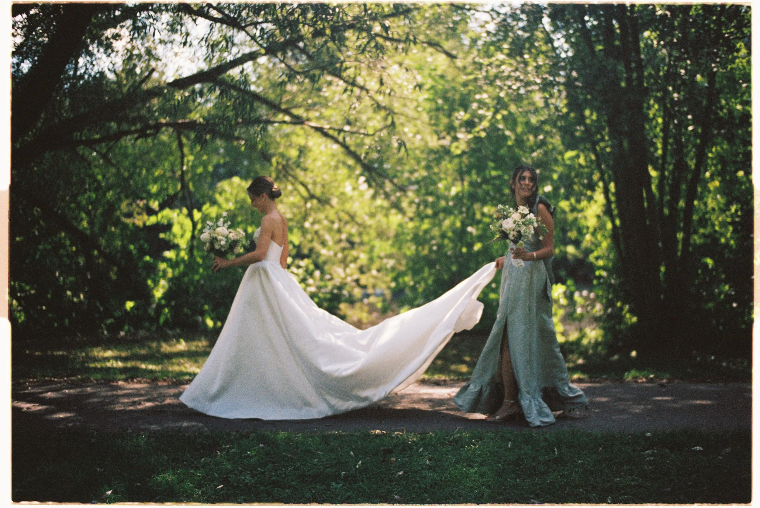 Two women in a park, one wearing a white wedding gown holding a bouquet, and the other wearing a long dress holding a bouquet, surrounded by lush green trees.