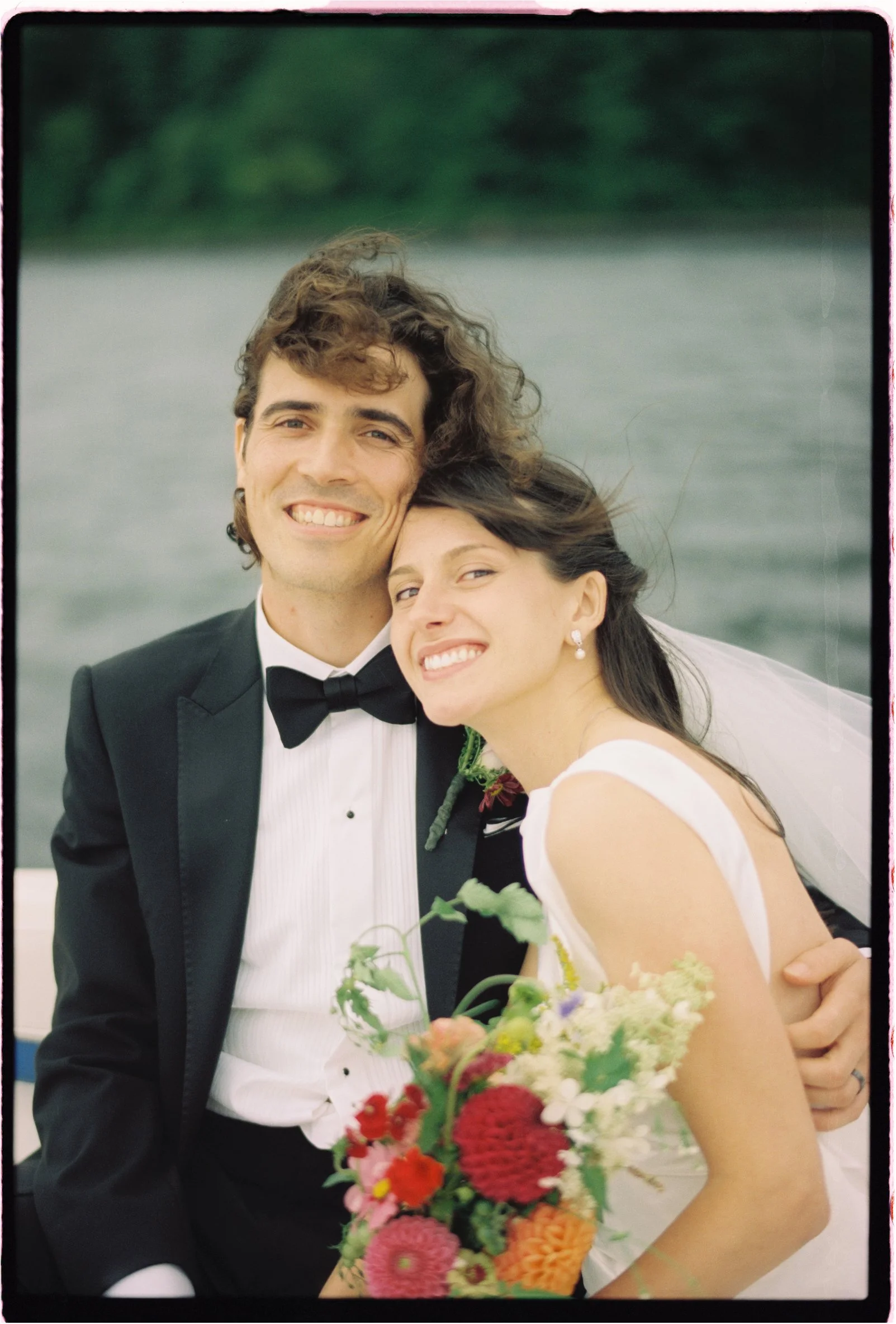 A smiling bride and groom in wedding attire embracing outdoors near a body of water, with the bride holding a colorful bouquet of flowers.