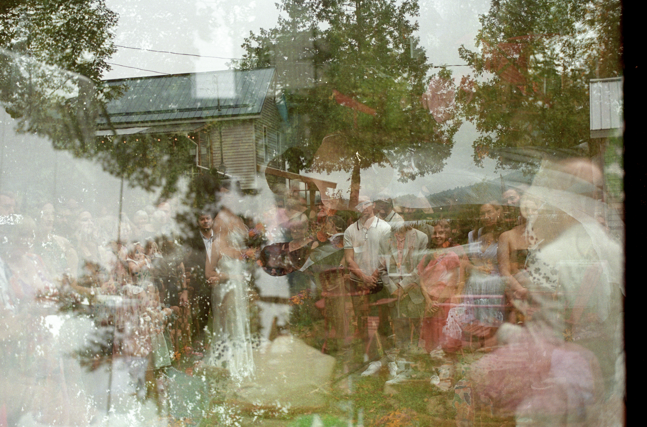 Multiple people gathered outdoors at a sunny event, with a building, trees, and a cloudy sky reflected in the glass, creating a layered, double exposure effect.
