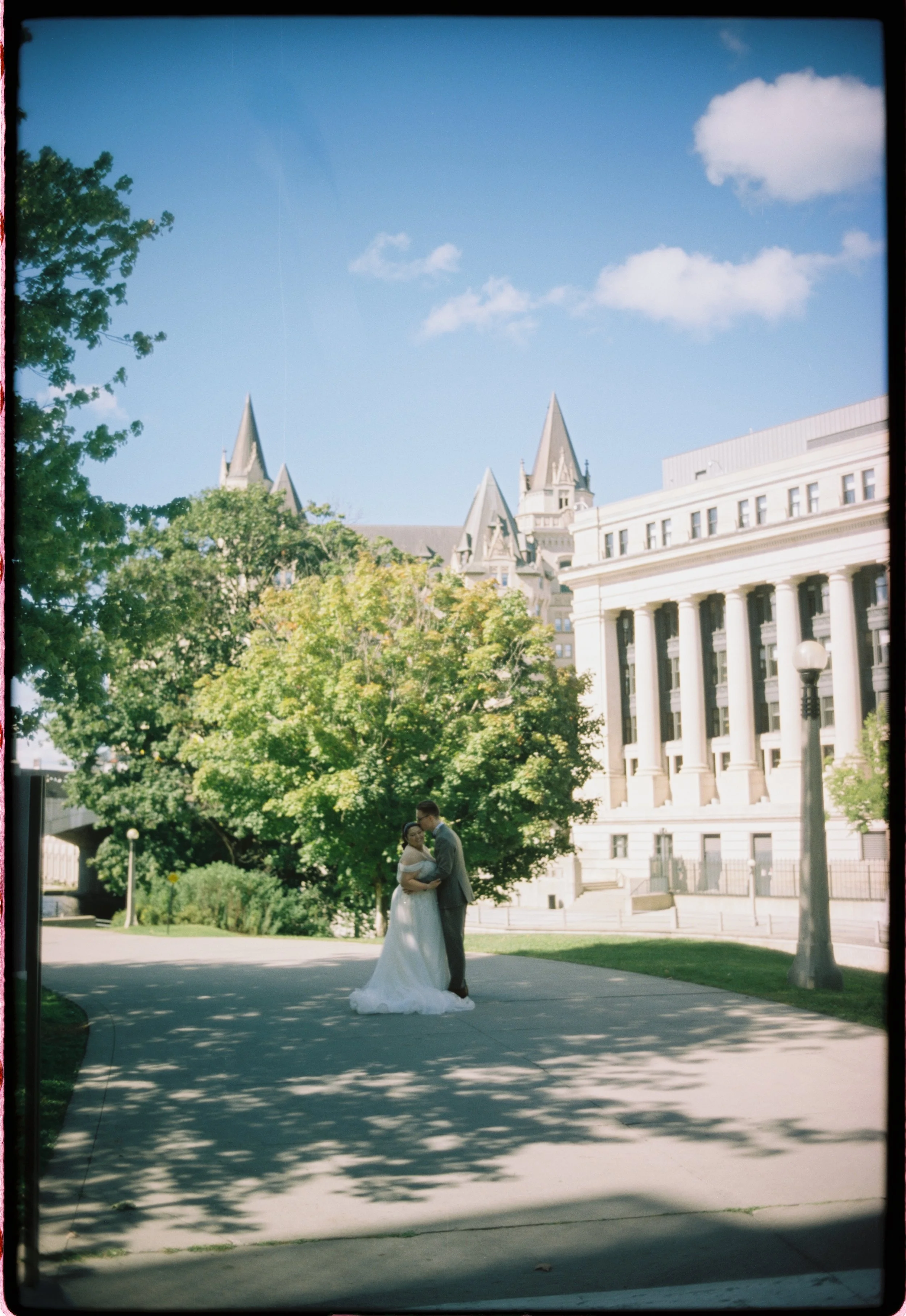 A bride and groom stand close together on a paved walkway in front of a large building with tall columns on a sunny day, with trees and a blue sky with clouds in the background.
