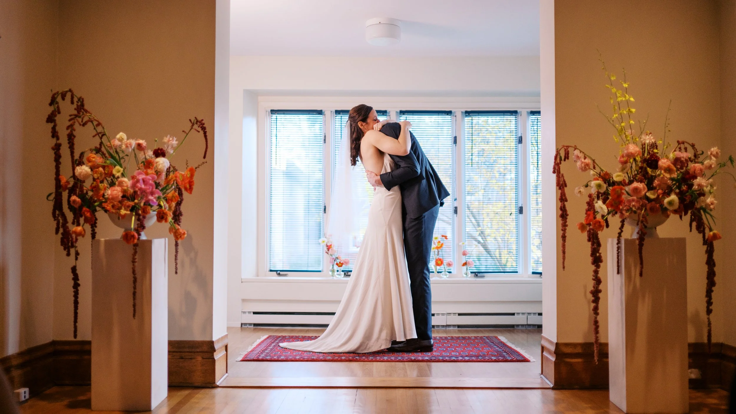 A bride and groom sharing a kiss in a bright room with large windows, decorated with colorful flowers in vases.