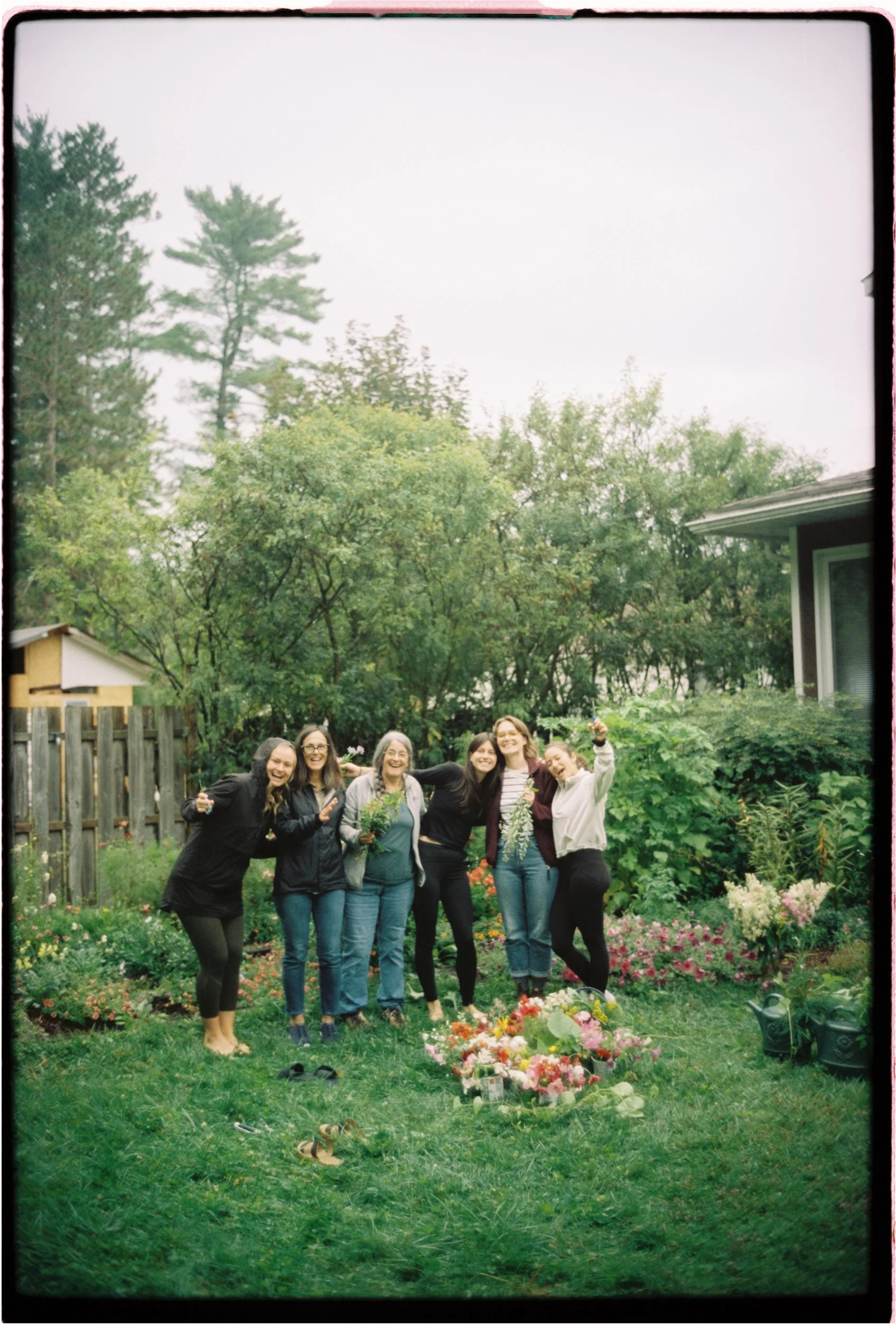 Group of six women celebrating in a garden with flowers, trees, and a wooden fence in the background.Ottawa Wedding Photo, film wedding, Kodak wedding, 35mm wedding
