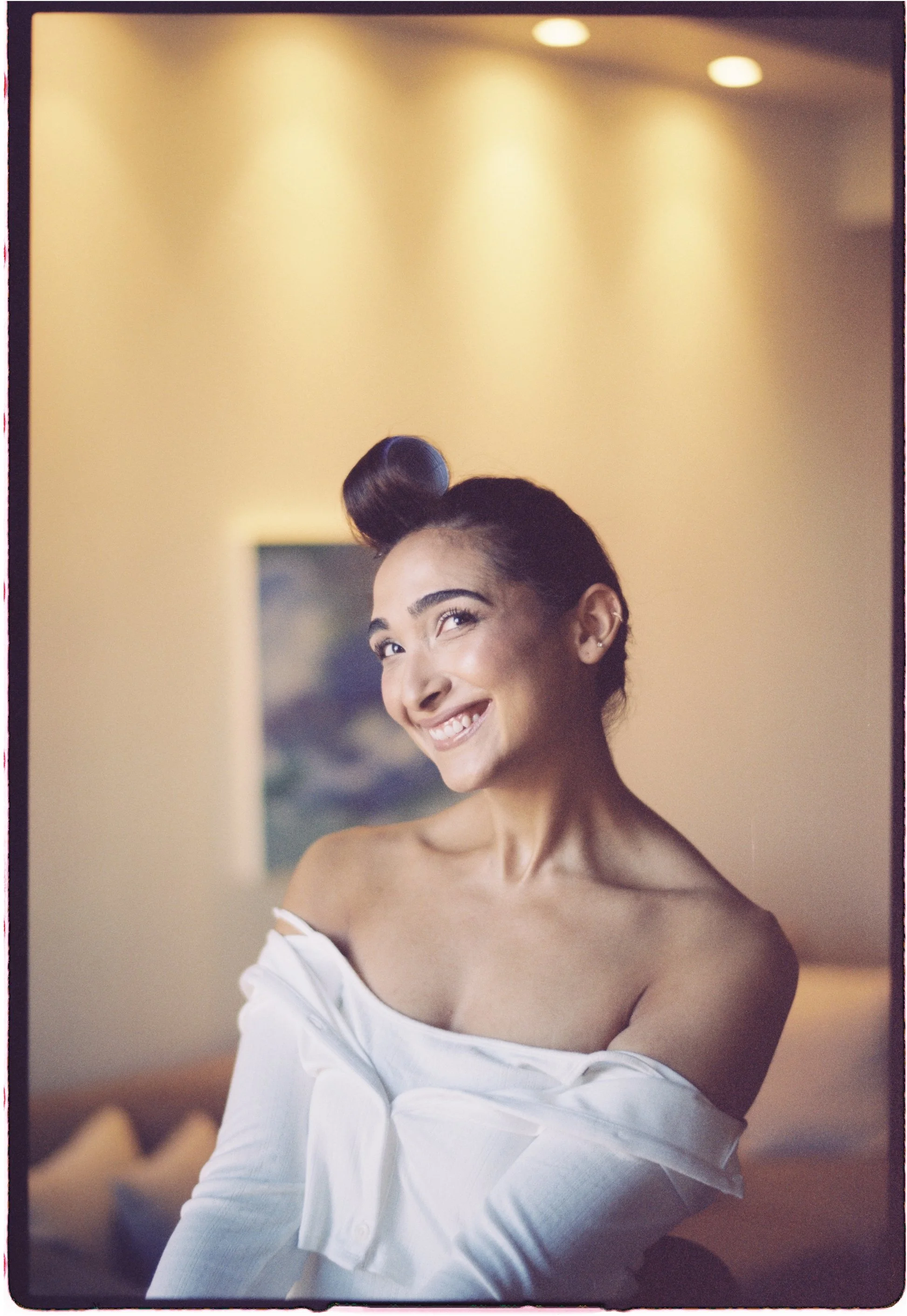 Ottawa Wedding Photo, film wedding, Kodak wedding, 35mm wedding A woman with dark hair styled in an updo and a white off-the-shoulder top, smiling and looking to the side in an indoor setting with warm lighting.