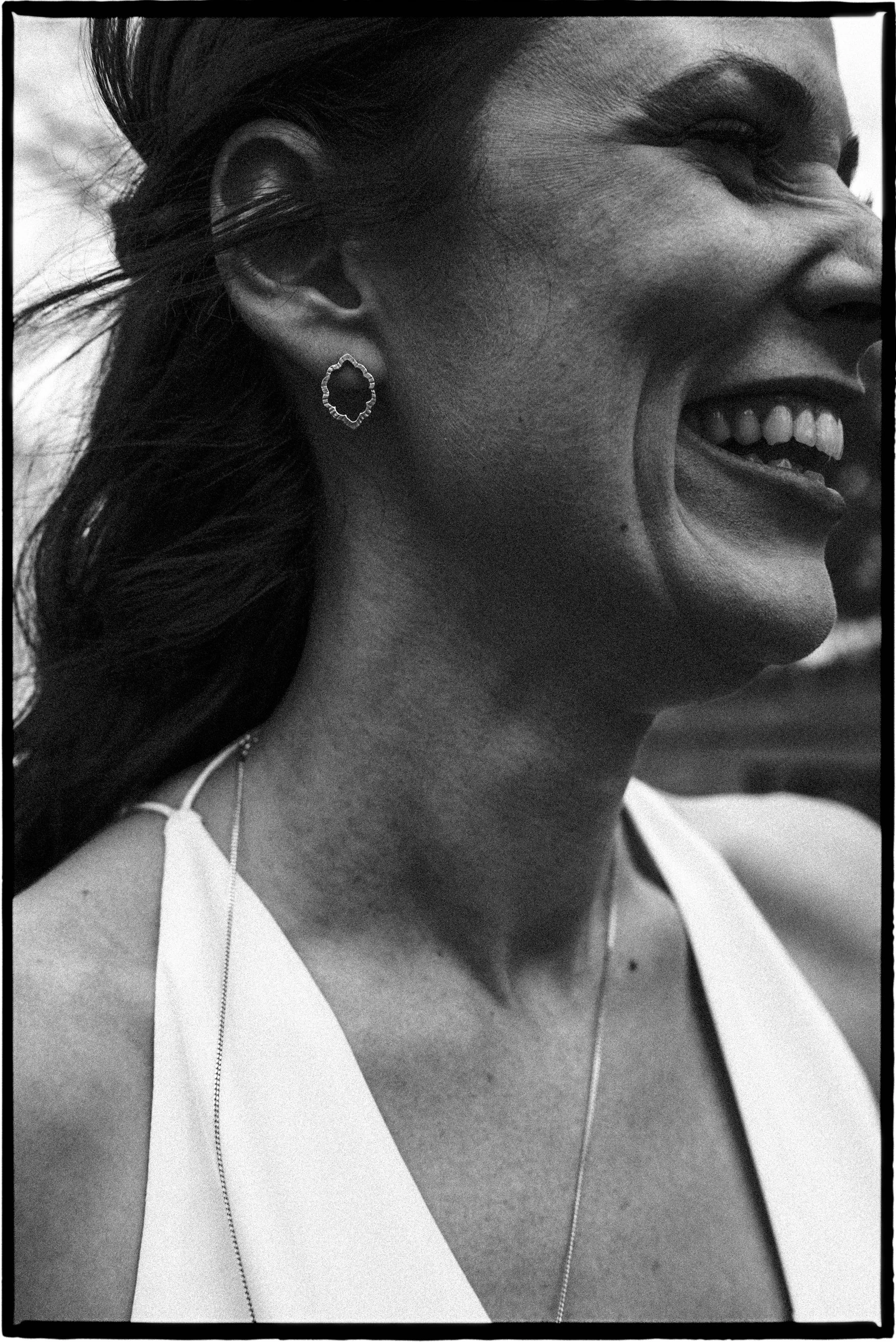 Close-up black and white photo of a woman laughing, showing her ear with a jewelry earring, part of her face, neck, and shoulder visible, wearing a sleeveless top and a necklace.
