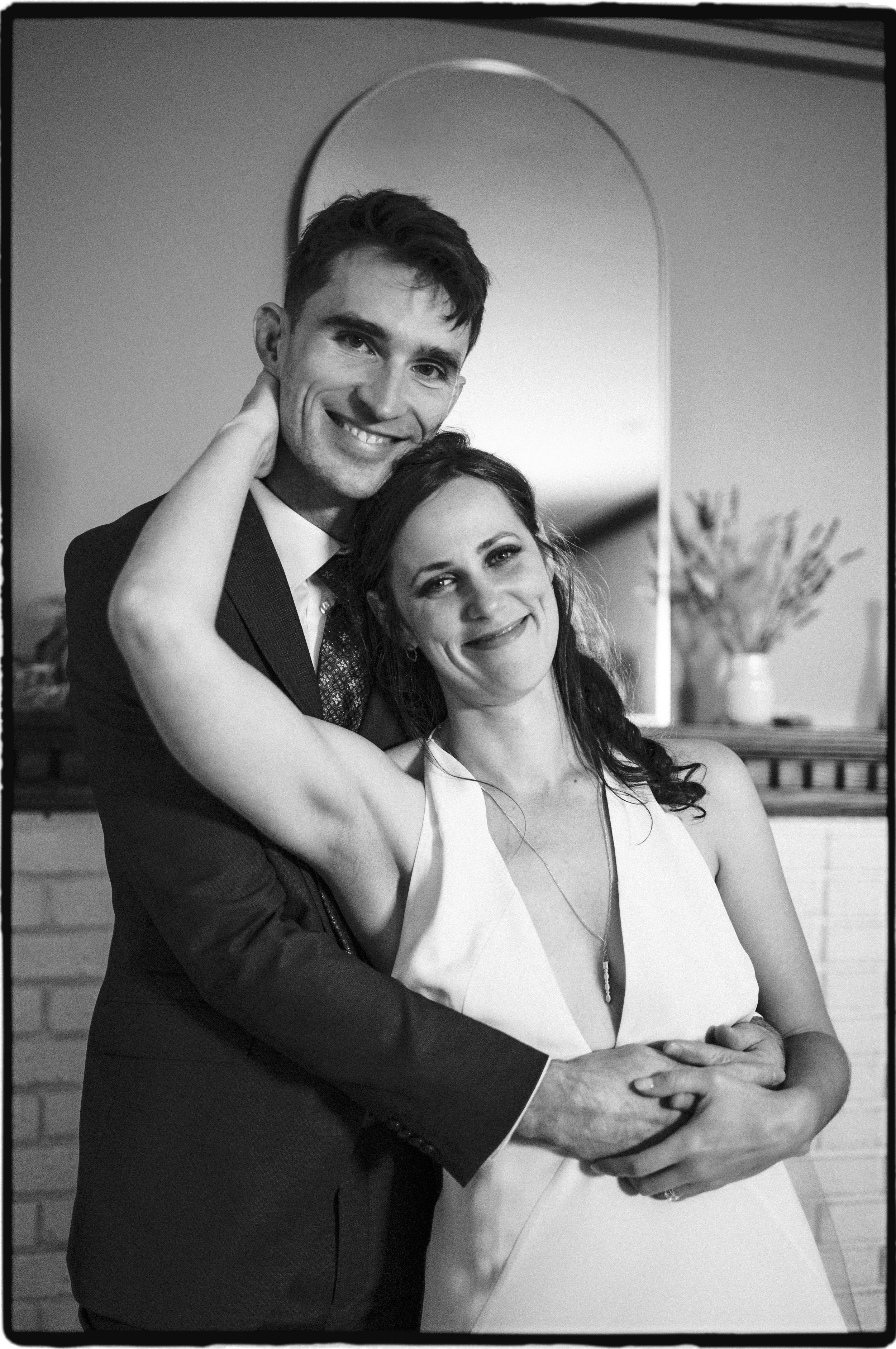 A black and white photo of a happy couple, a man in a suit and a woman in a sleeveless dress, smiling and posing together in front of a mirror.Ottawa Wedding Photo, film wedding, Kodak wedding, 35mm wedding
