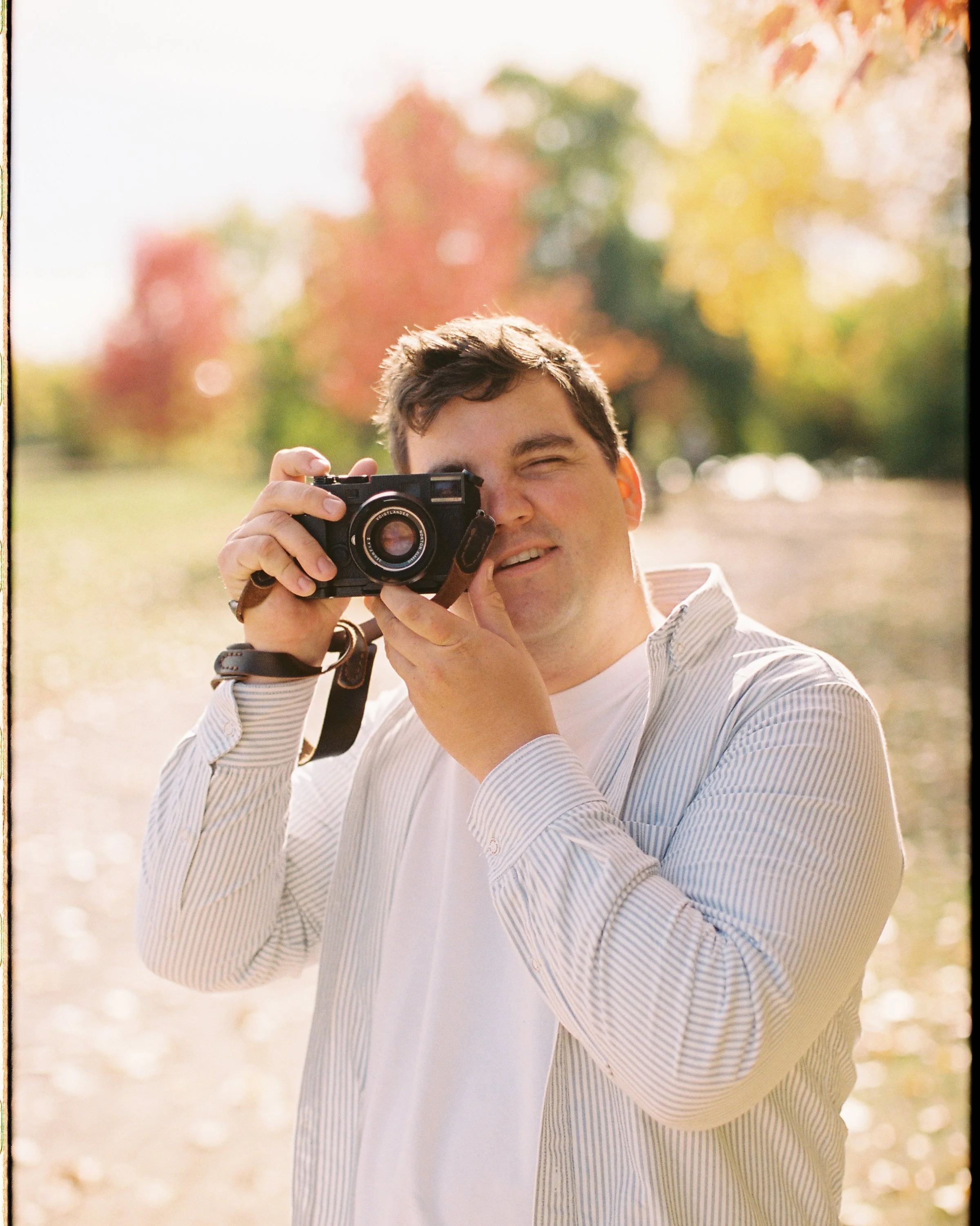 A young man in a light-colored, striped shirt, holding a camera to his face, outdoors with fall foliage and trees in the background.
