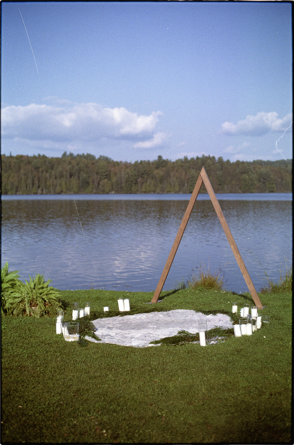 An outdoor memorial setup near a lake with a wooden A-frame structure, surrounded by candles and greenery, with a forest in the background under a blue sky with clouds.