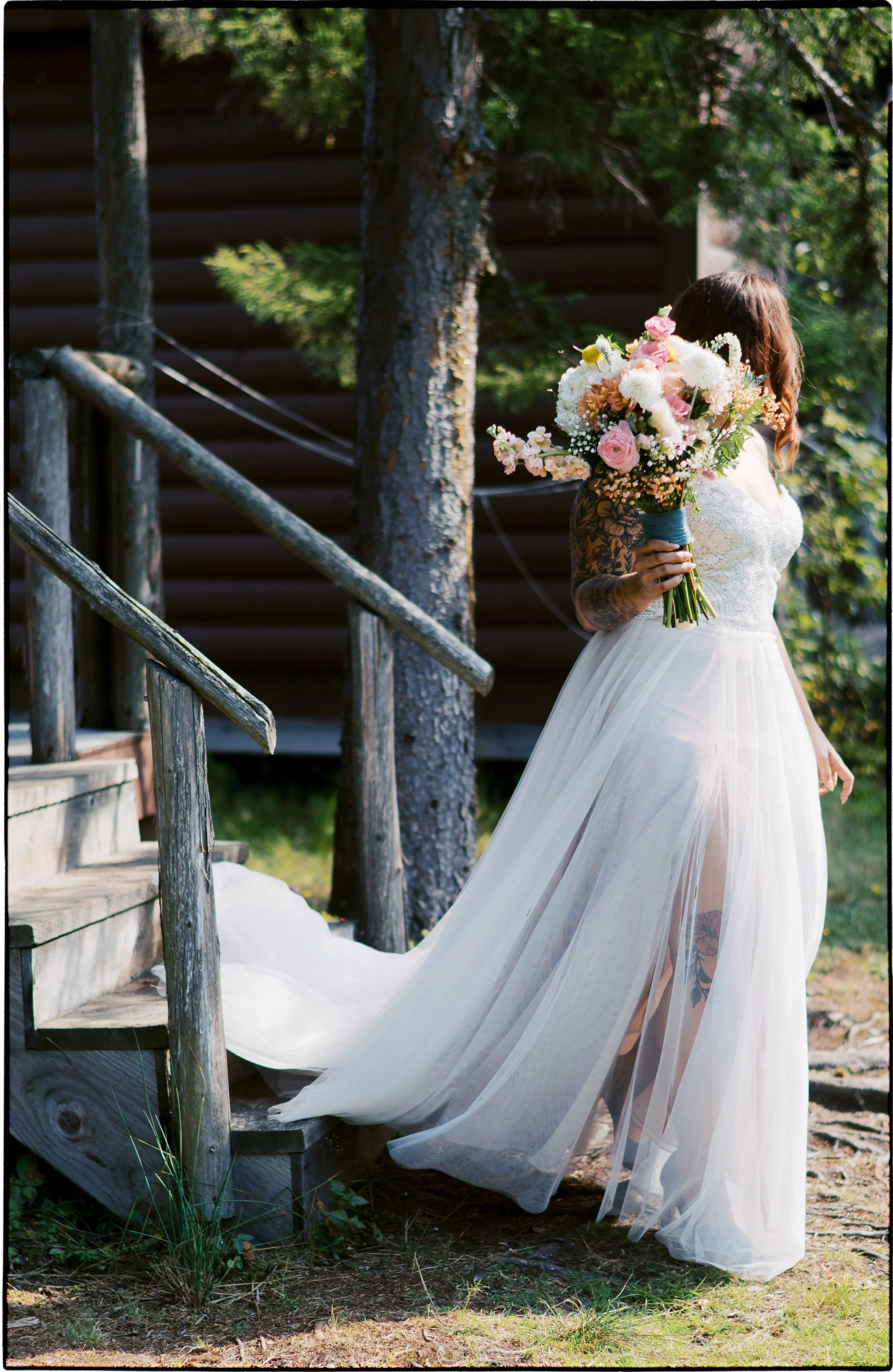 A bride in a white wedding dress holding a colorful bouquet of flowers, standing outdoors near a wooden staircase and trees.