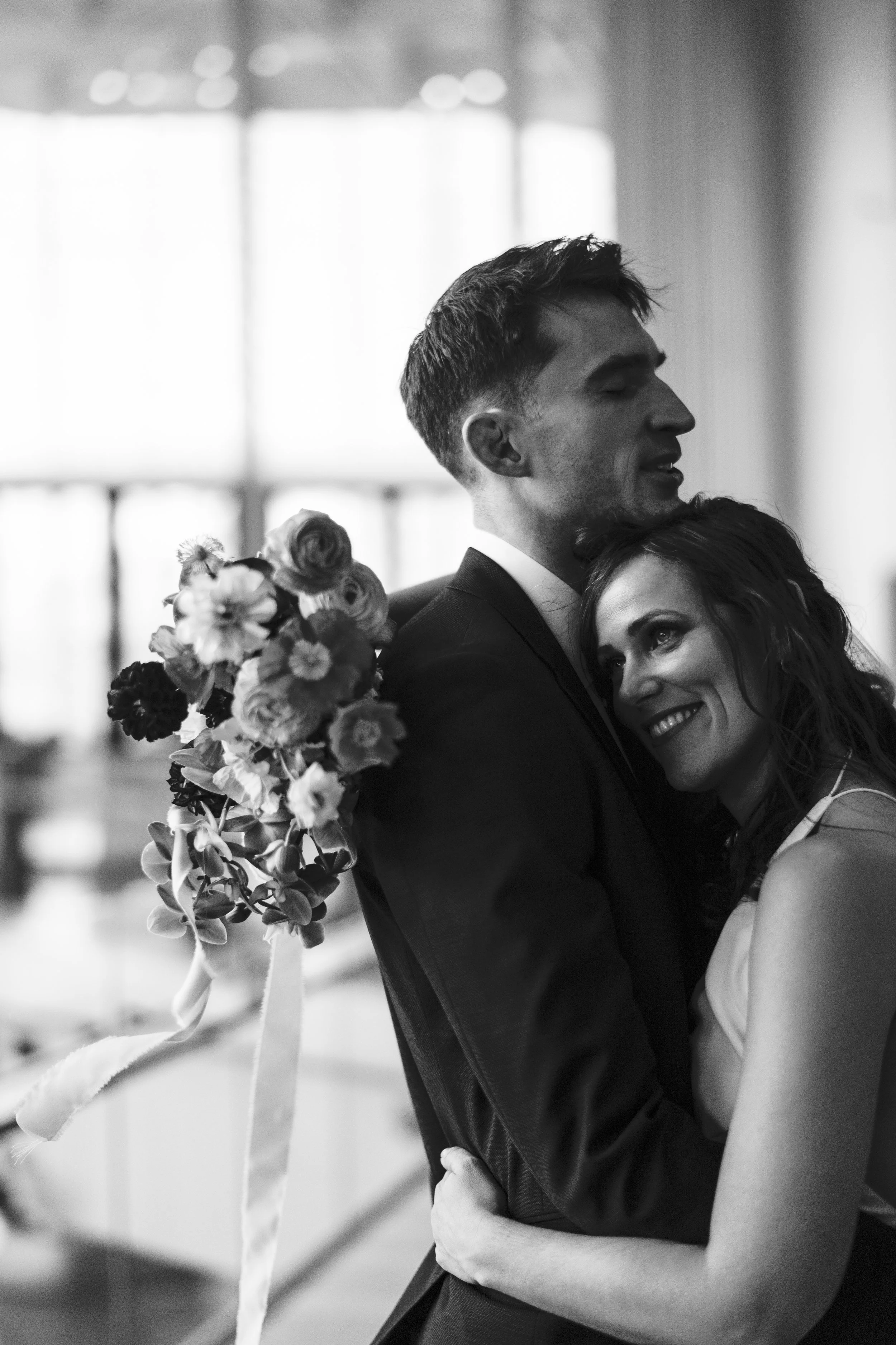 A black-and-white photograph of a couple embracing, with the woman smiling and looking at the camera while holding a bouquet of flowers, and the man with eyes closed, in formal attire, likely at a wedding or special event.