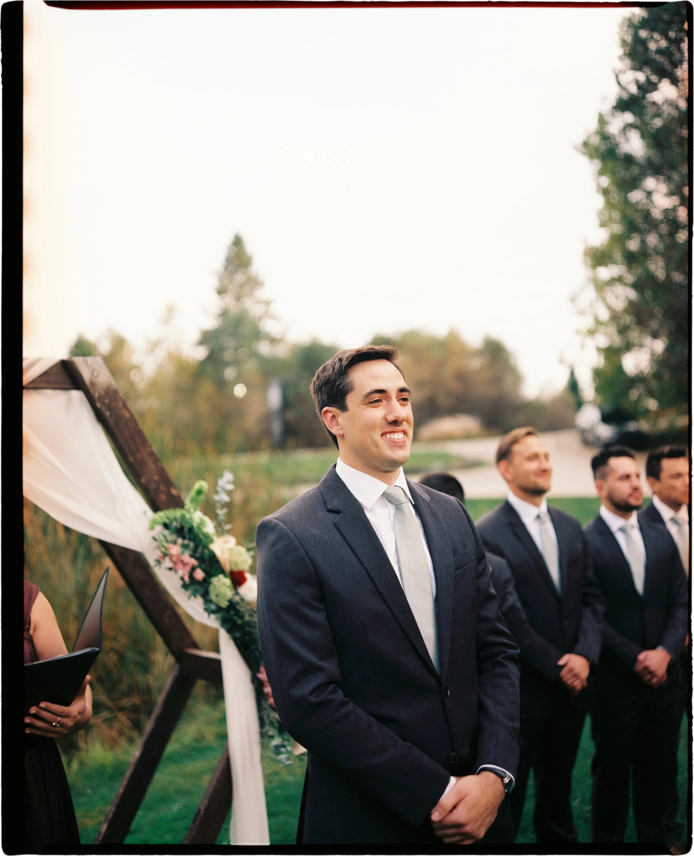 Ottawa Wedding Photo, film wedding, Kodak wedding, 35mm wedding groom in a dark suit smiles during an outdoor wedding ceremony, with groomsmen standing behind him.