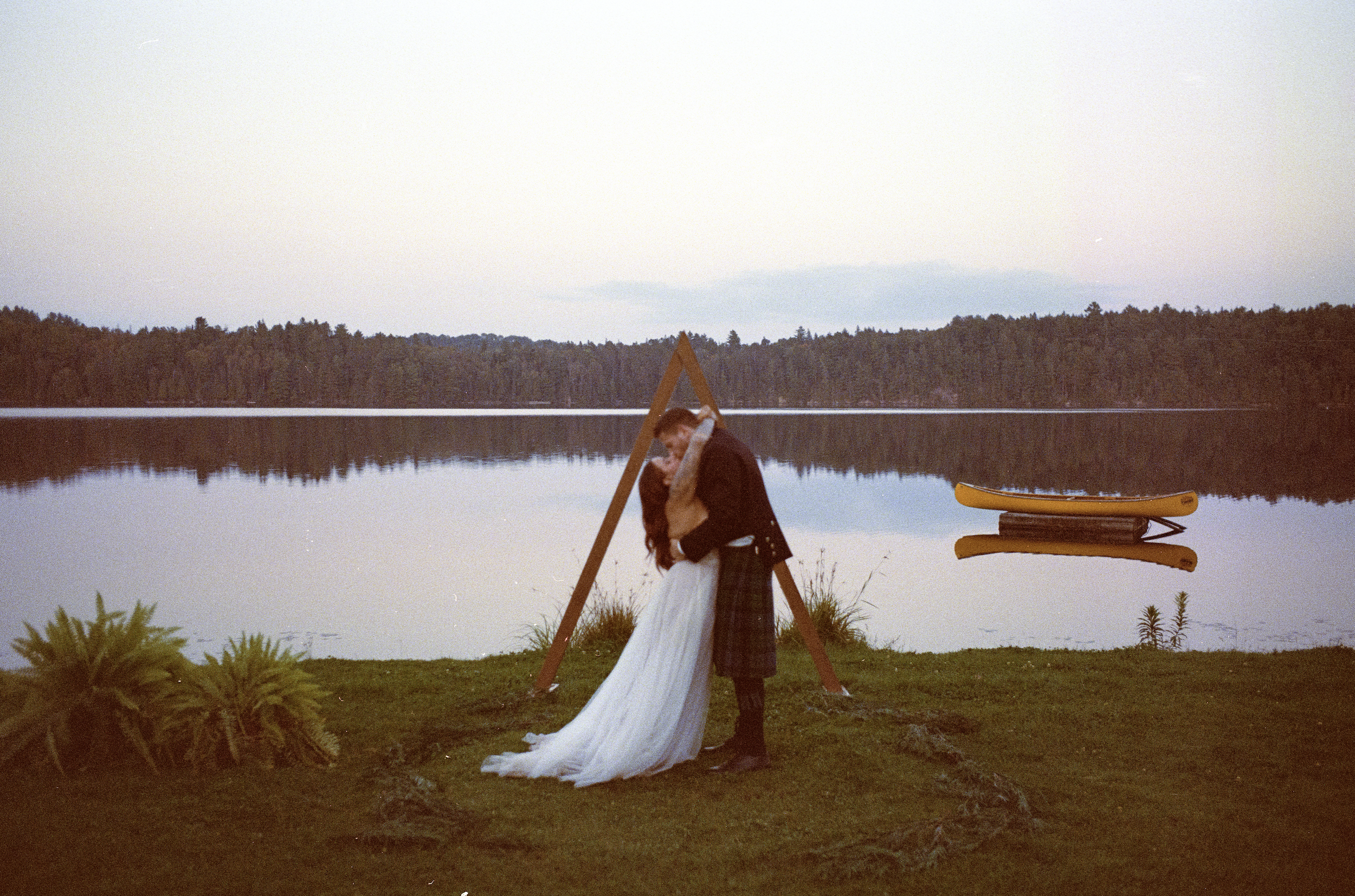 A couple embracing and kissing by a lake during sunset, with a wooden triangular structure and a yellow kayak on the shore.