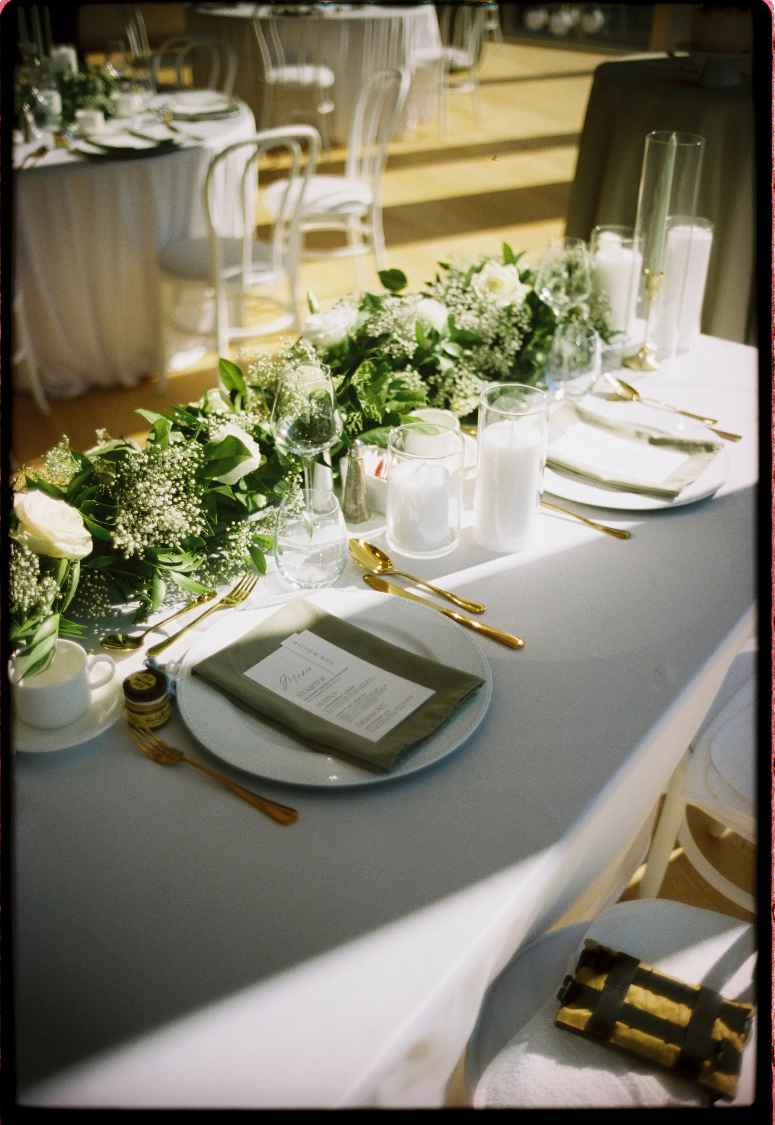 Elegant wedding reception table decorated with white flowers, greenery, candles, and gold cutlery, set with plates, glasses, and menus.