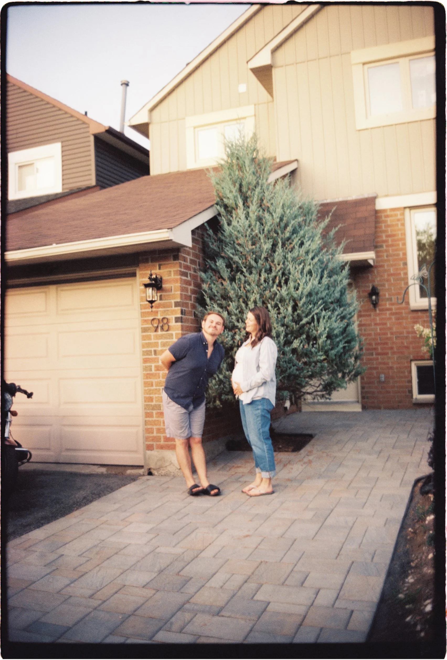 Two people, a man and a woman, standing outside in front of a house with a tall evergreen tree. The man is wearing a navy shirt and shorts, and the woman is pregnant, wearing a white jacket and jeans. The house has a brick exterior, a garage door, an