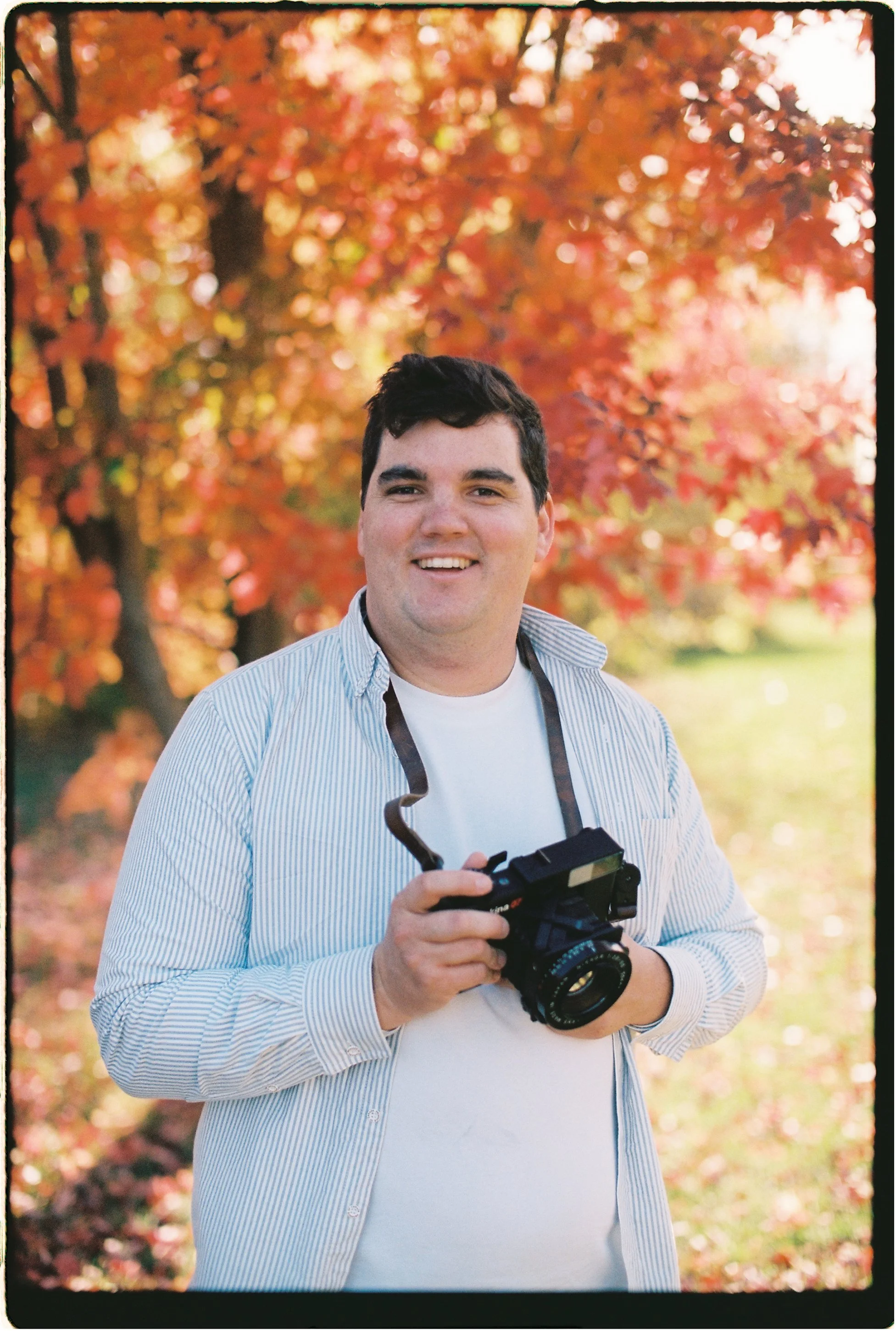 A man smiling outdoors in front of colorful autumn trees, holding a camera.