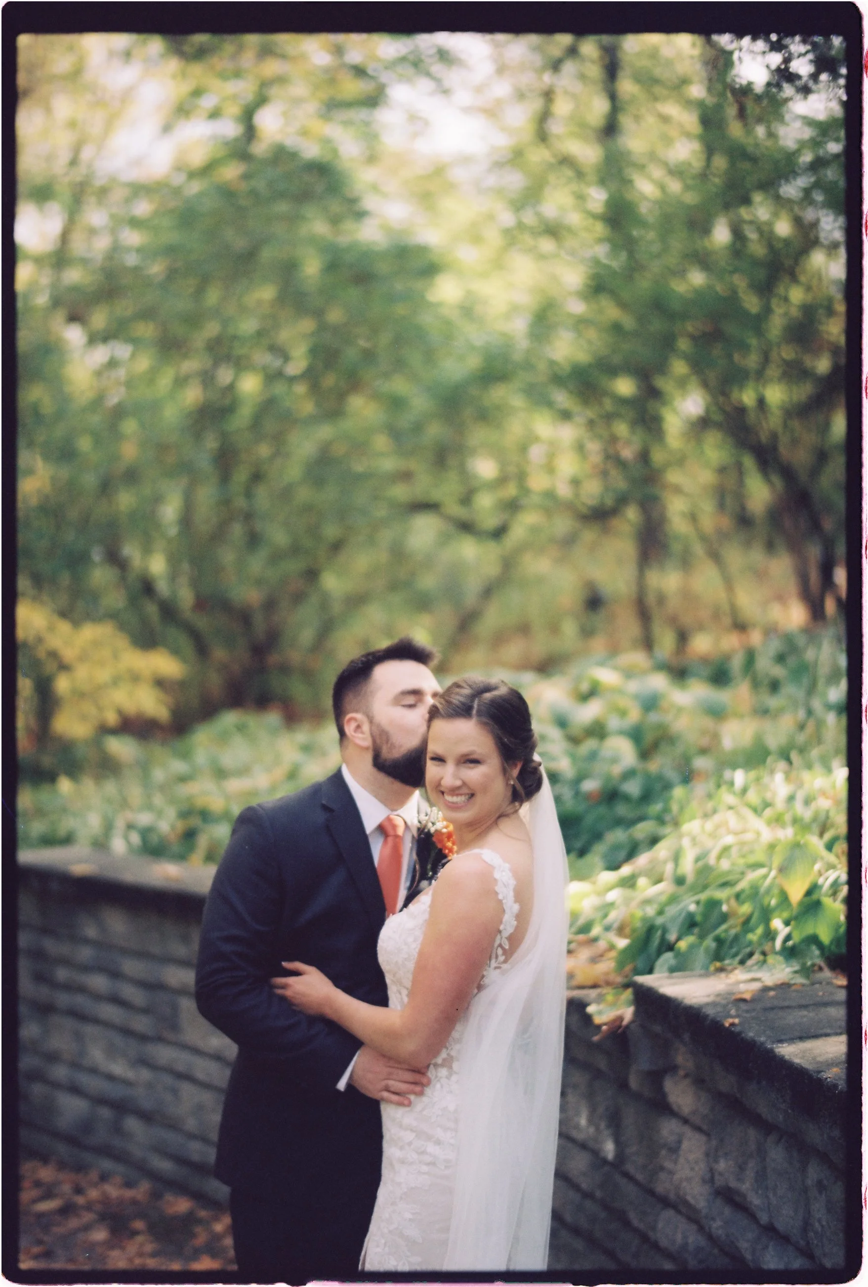 A newlywed couple standing outdoors on a stone wall, with the groom kissing the bride on the forehead and the bride smiling happily. The background features green trees and foliage.