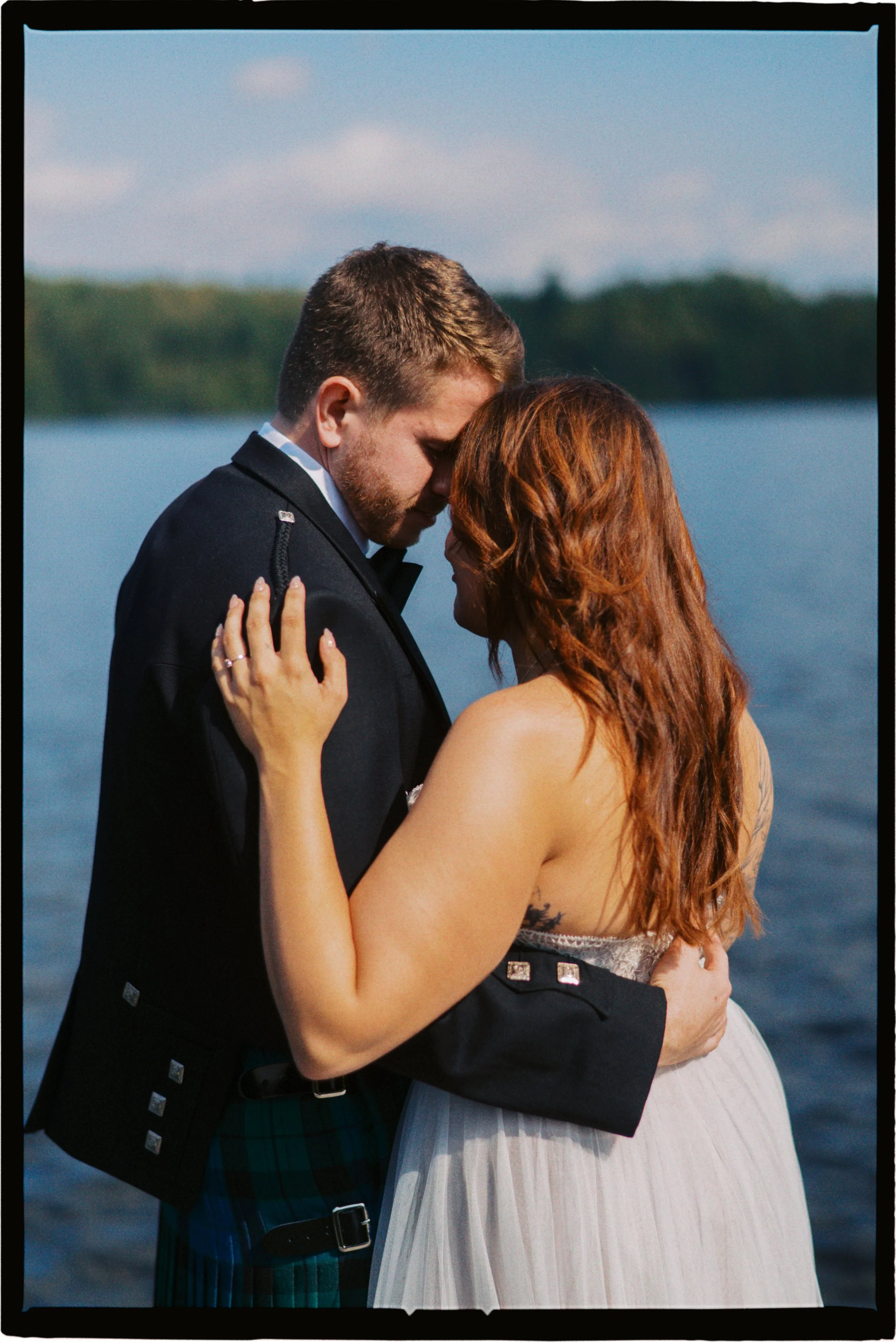 A couple embraces by the water, with the man dressed in a formal black jacket and the woman in a strapless dress, both with their foreheads touching.