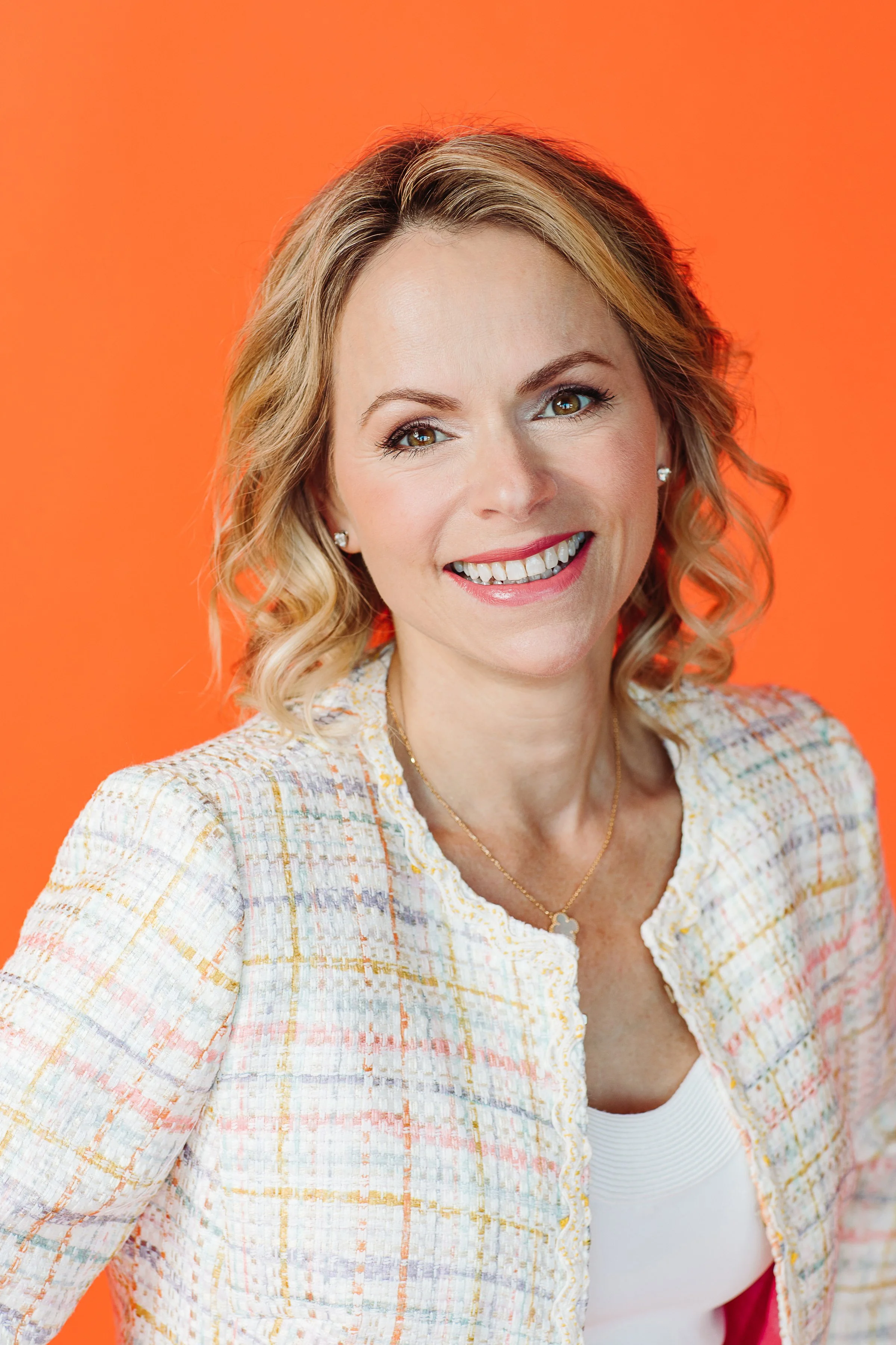 Robin Ducharme with shoulder-length blonde wavy hair wearing a colorful tweed jacket and a white top against an orange background.