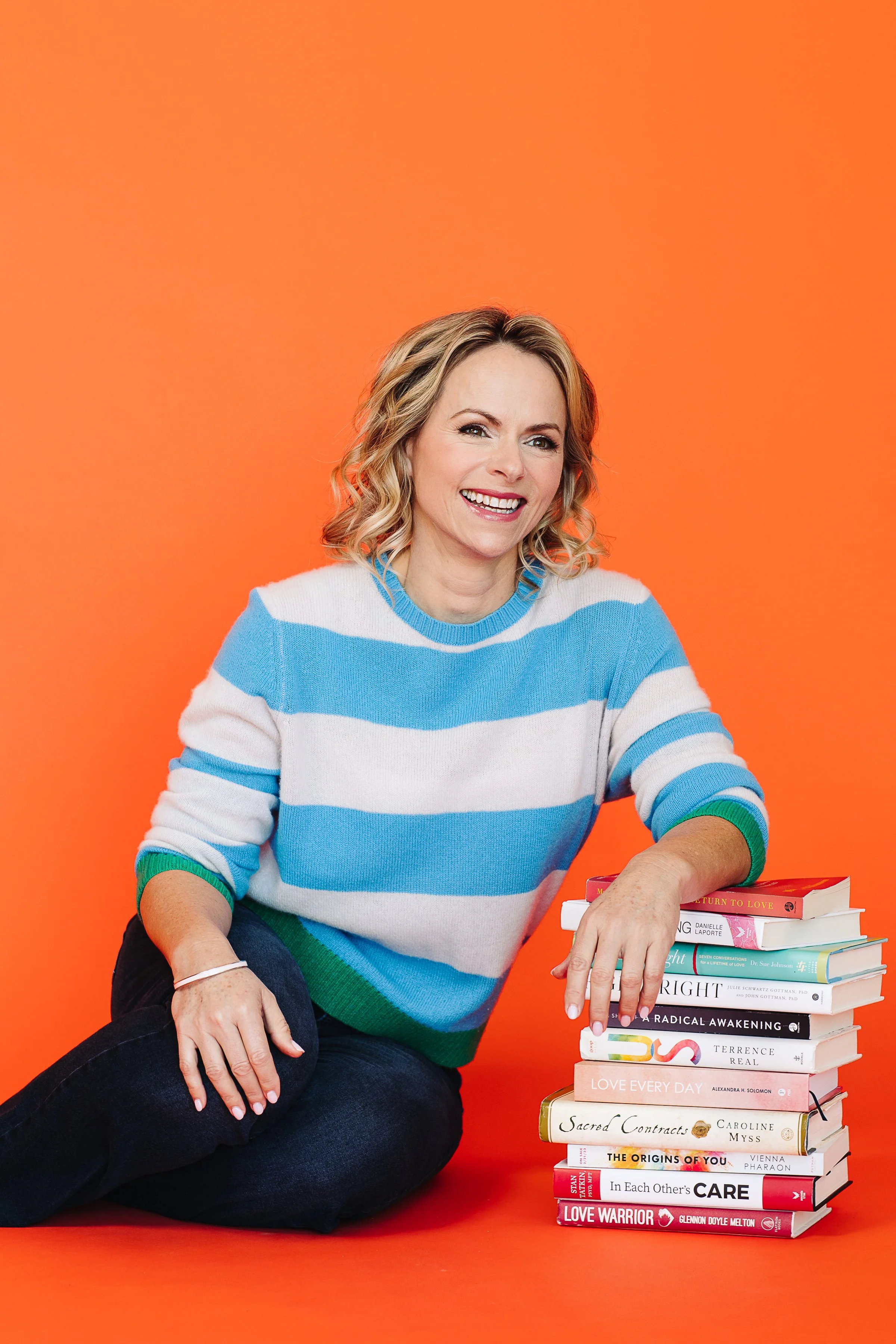 A woman with blonde, wavy hair smiling and sitting next to a stack of books against an orange background, wearing a blue and white striped sweater and dark pants.