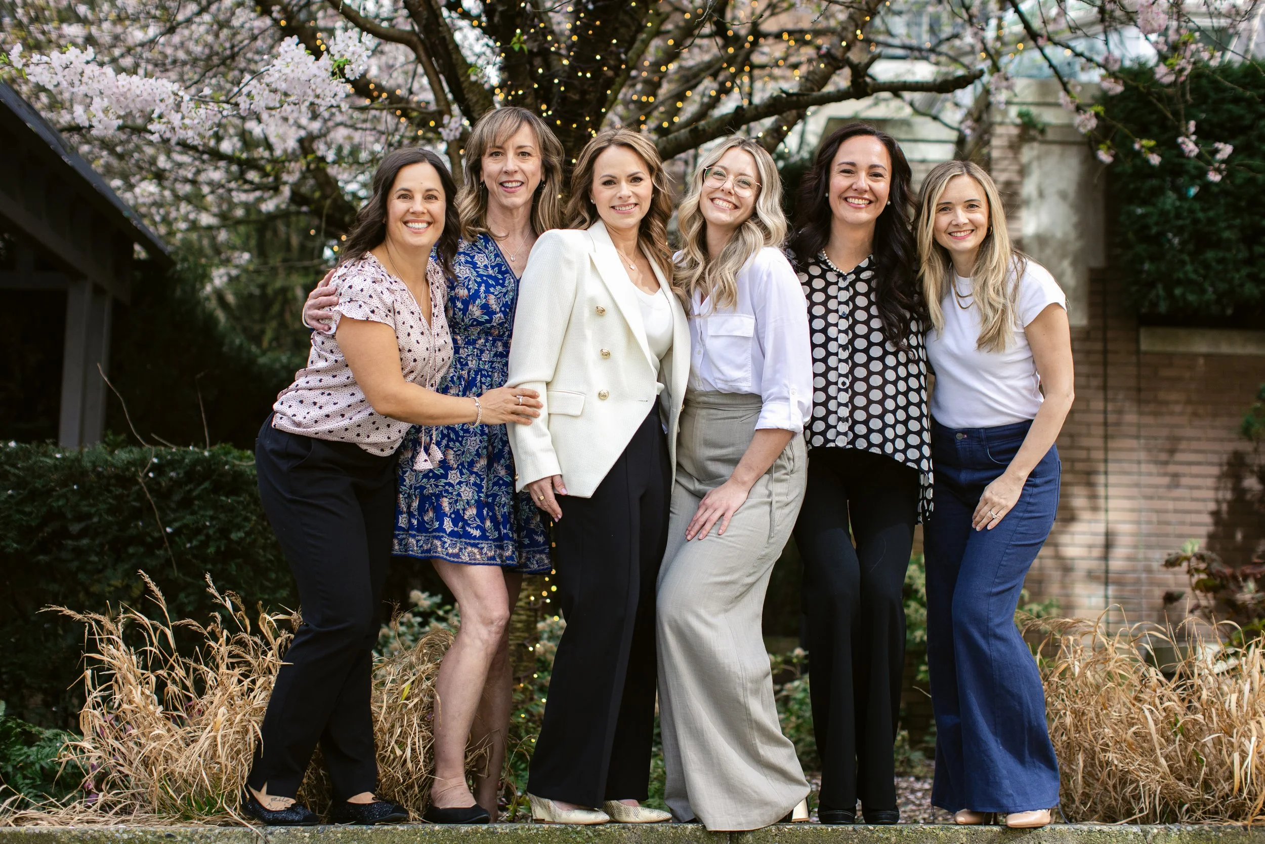 Group of seven women standing outdoors in front of a blossoming tree, enjoying a sunny day and dressed in semi-formal attire.