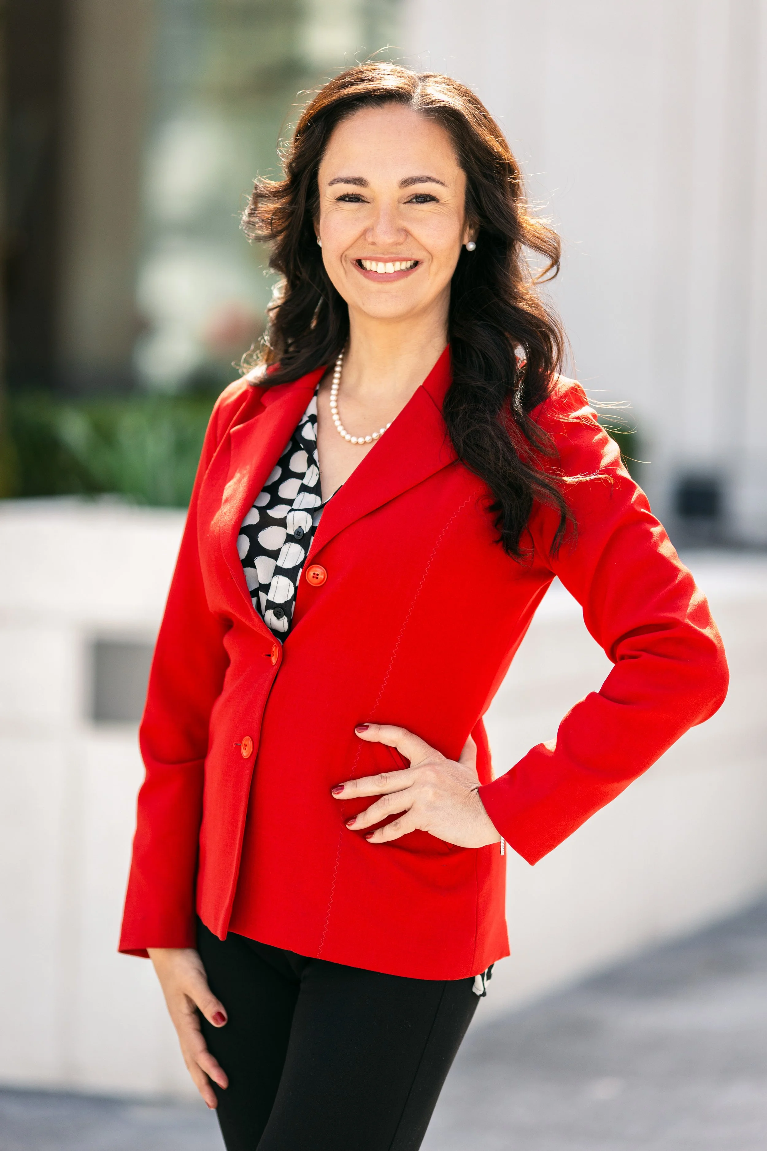 A smiling woman with wavy brown hair wearing a red blazer, black and white patterned blouse, black pants, pearl necklace, and pearl earrings standing outdoors during daytime.
