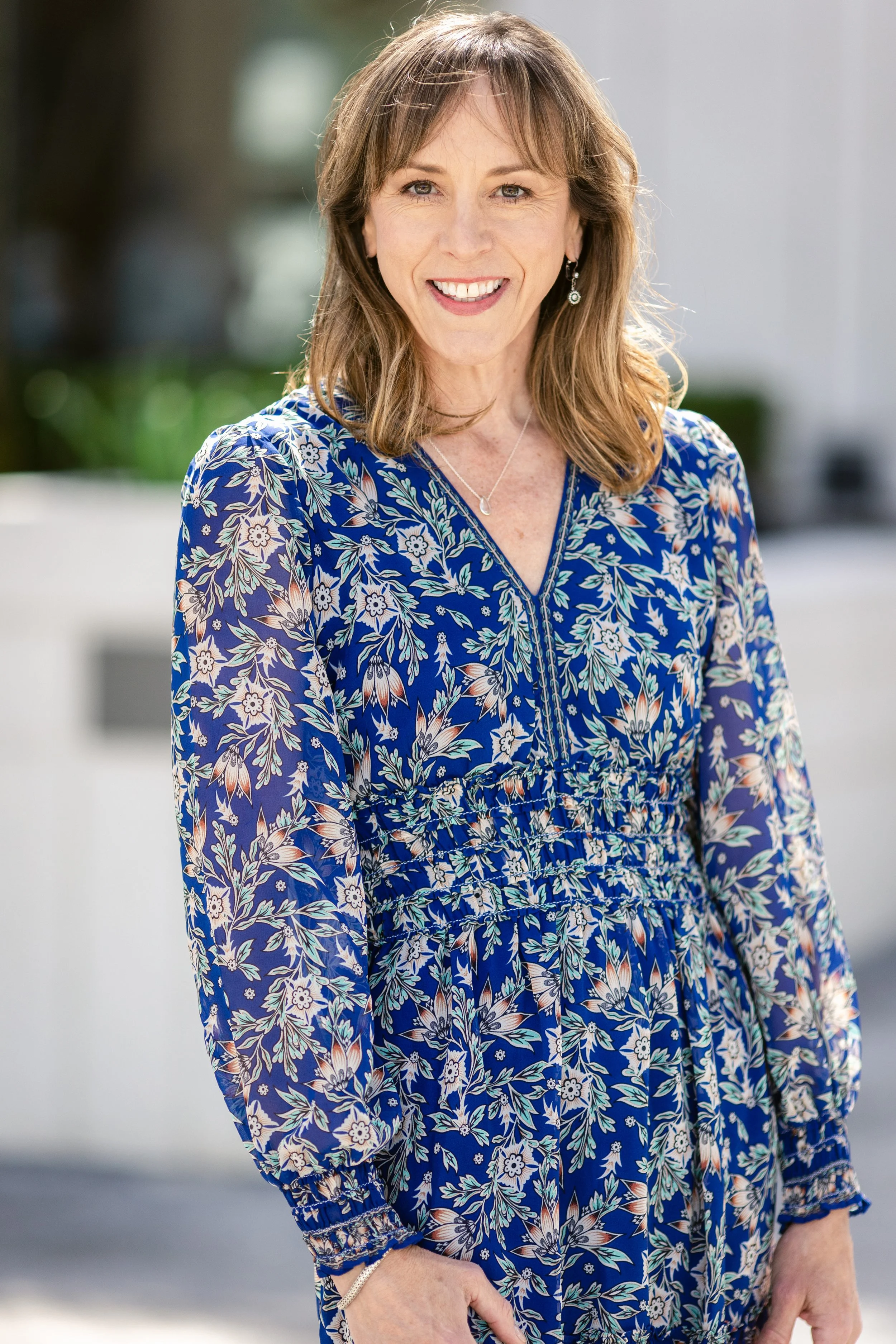 A woman with shoulder-length light brown hair, smiling, wearing a blue floral dress with long sleeves and a V-neck.