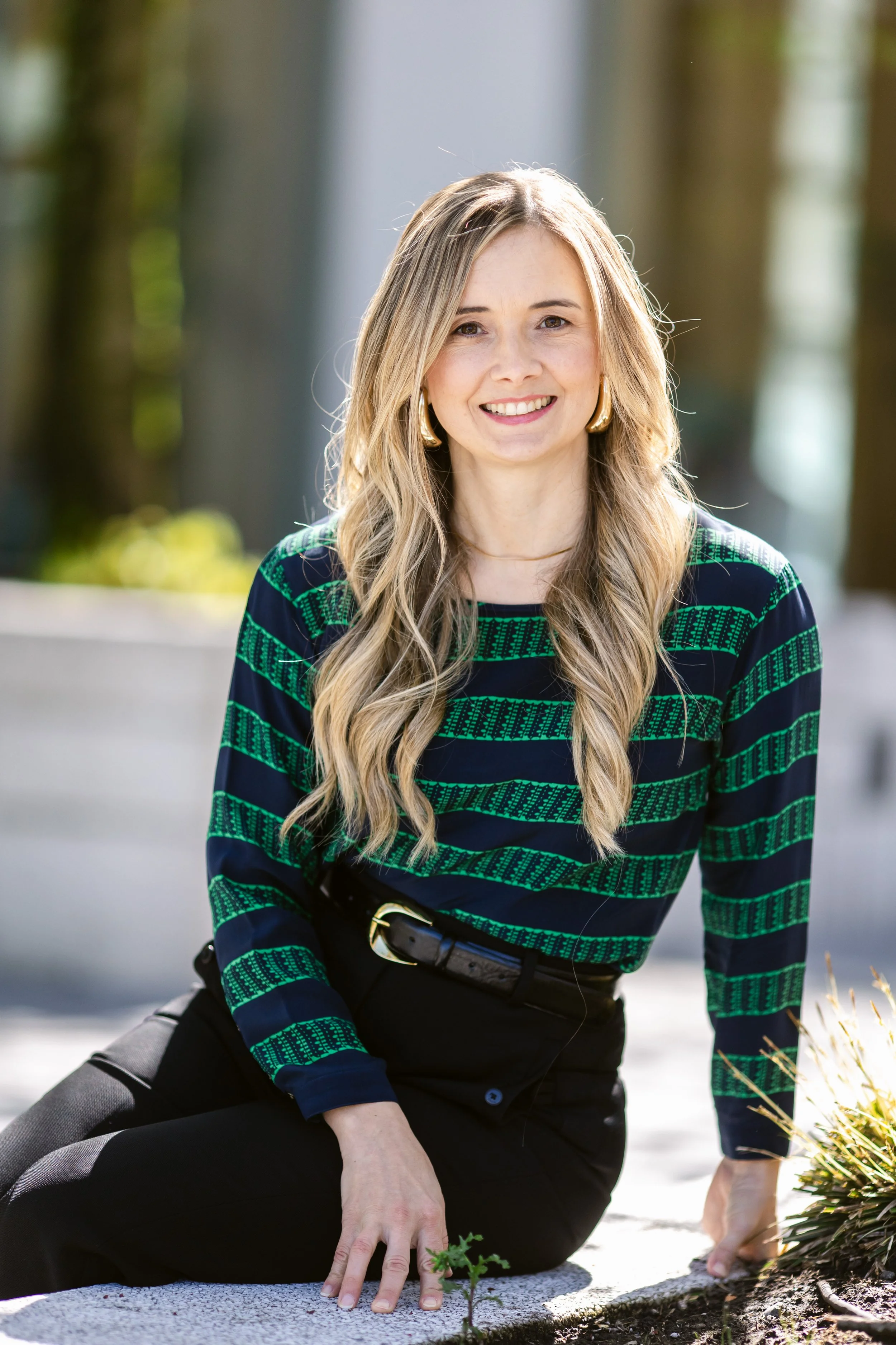 A photo of Kate Muker sitting outside, smiling and wearing a green and navy striped sweater with a belt and black pants.