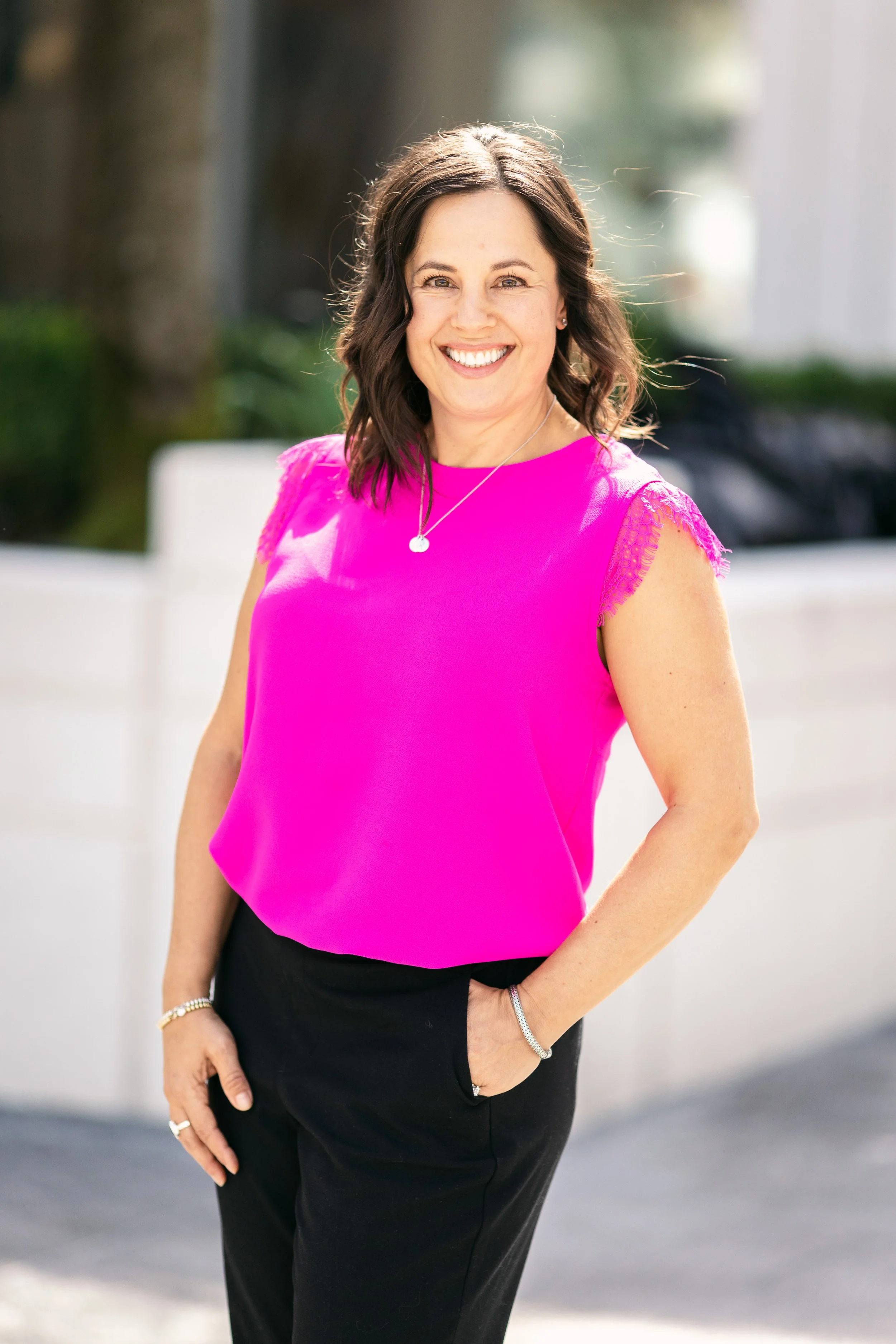 A woman with shoulder-length brown hair smiling, wearing a bright pink sleeveless top and black pants, standing outdoors.