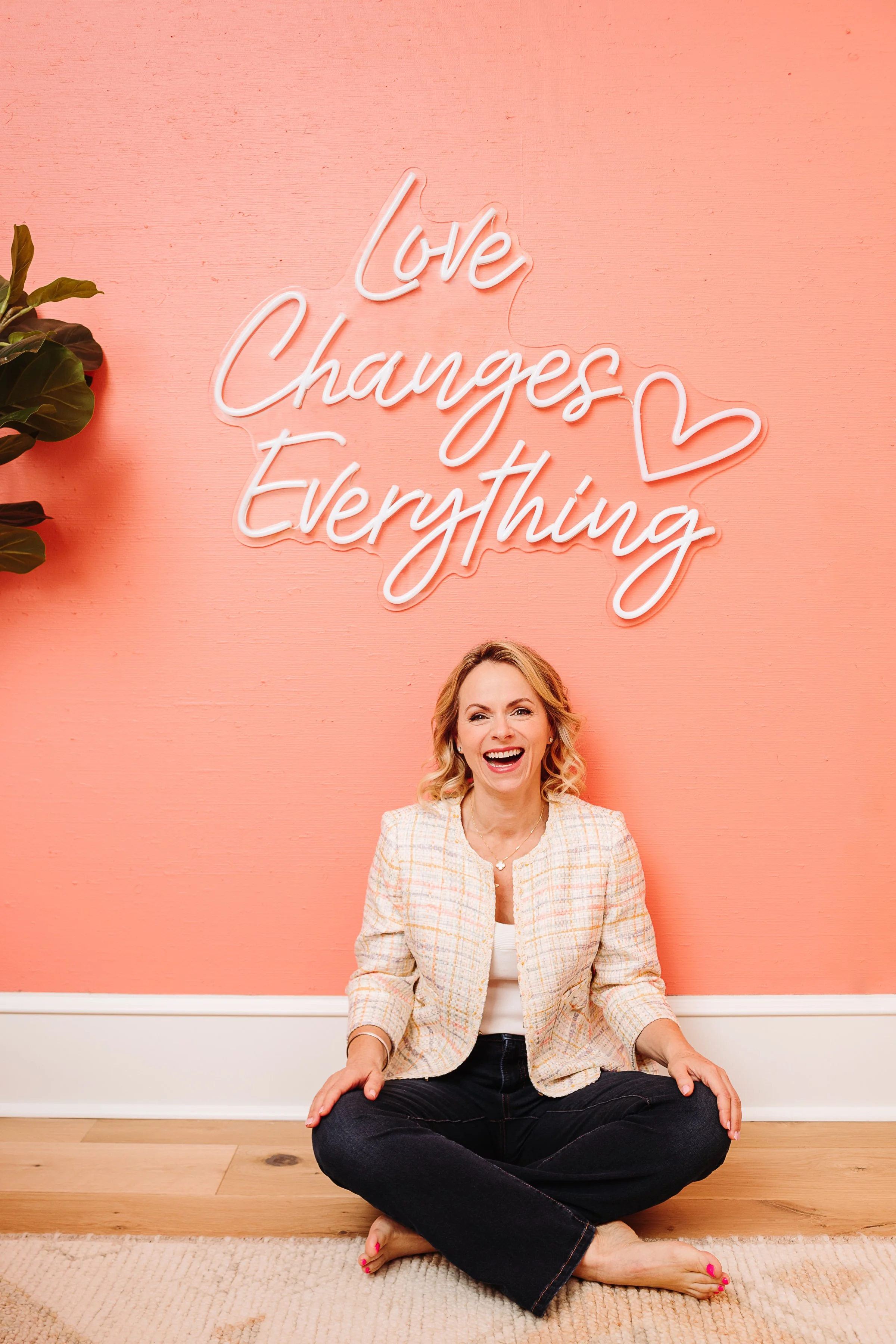A woman sitting cross-legged on the floor laughing in front of a pink wall with a neon sign that says 'Love Changes Everything' and a heart outline.