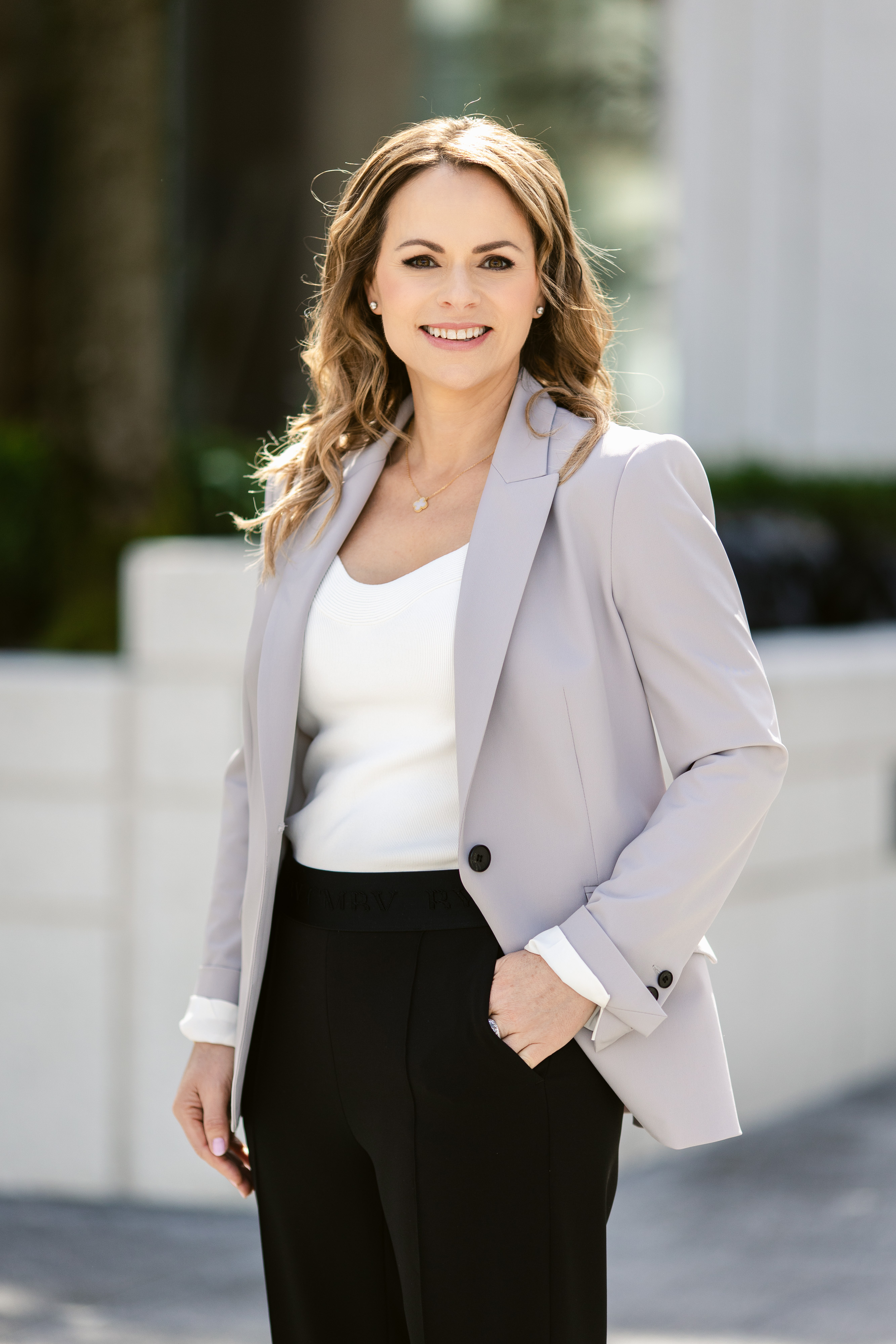 A woman with light brown, wavy hair smiling outdoors, wearing a gray blazer over a white top and black pants.