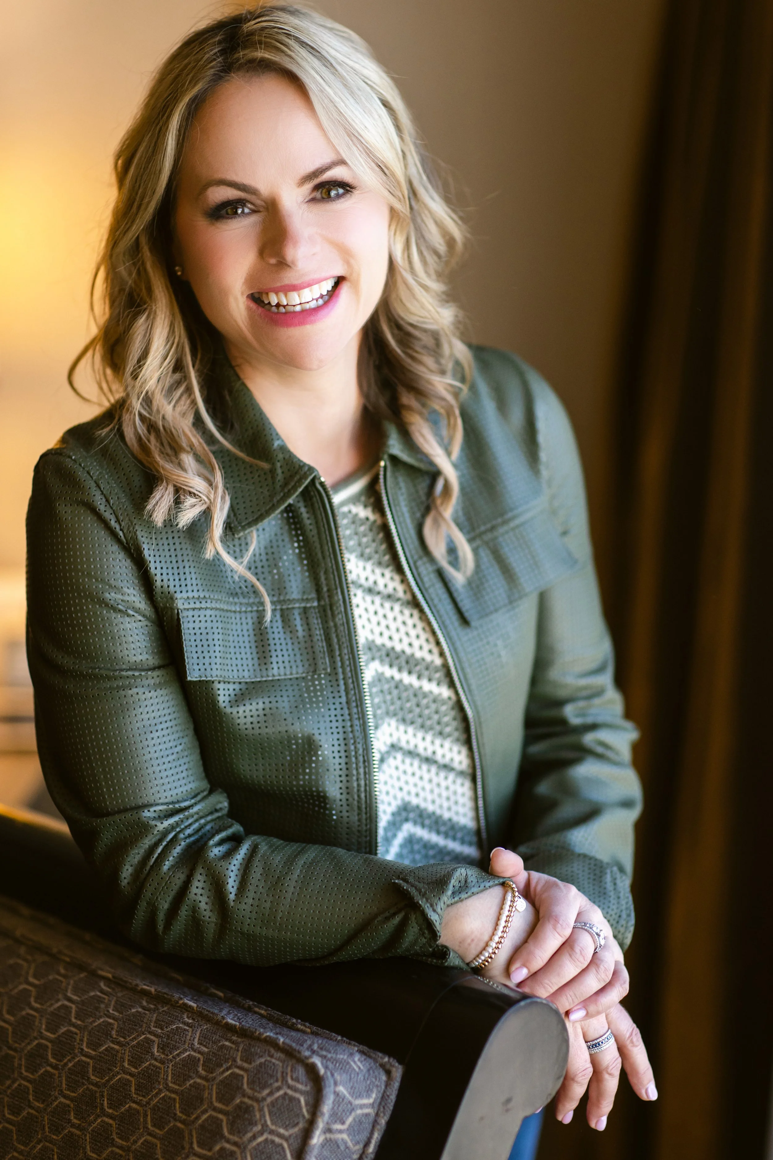 A woman with blonde wavy hair smiling, wearing a dark green jacket and patterned top, standing indoors with warm lighting.