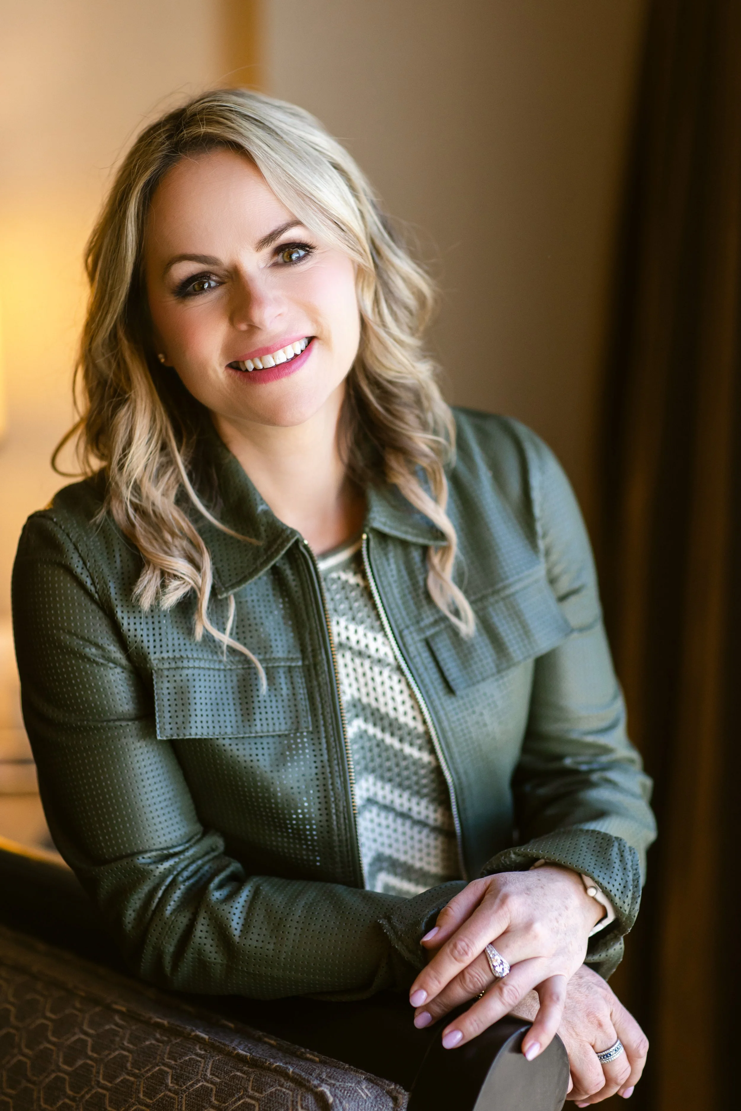 A woman with blonde hair smiling and sitting indoors, wearing a green jacket and patterned shirt.