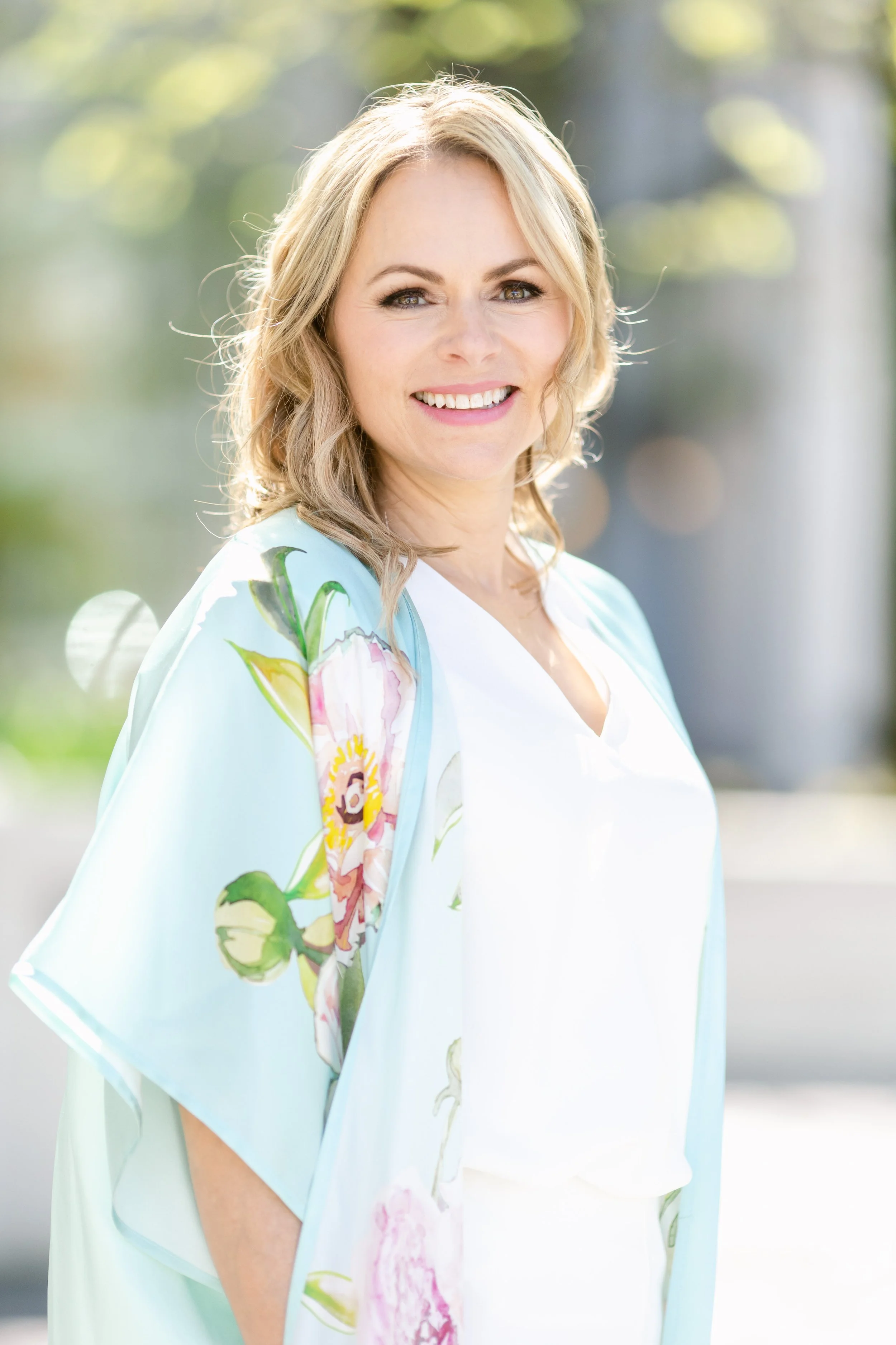 A woman with blonde hair and blue eyes smiling outdoors, wearing a white top and a light blue embroidered floral shawl.