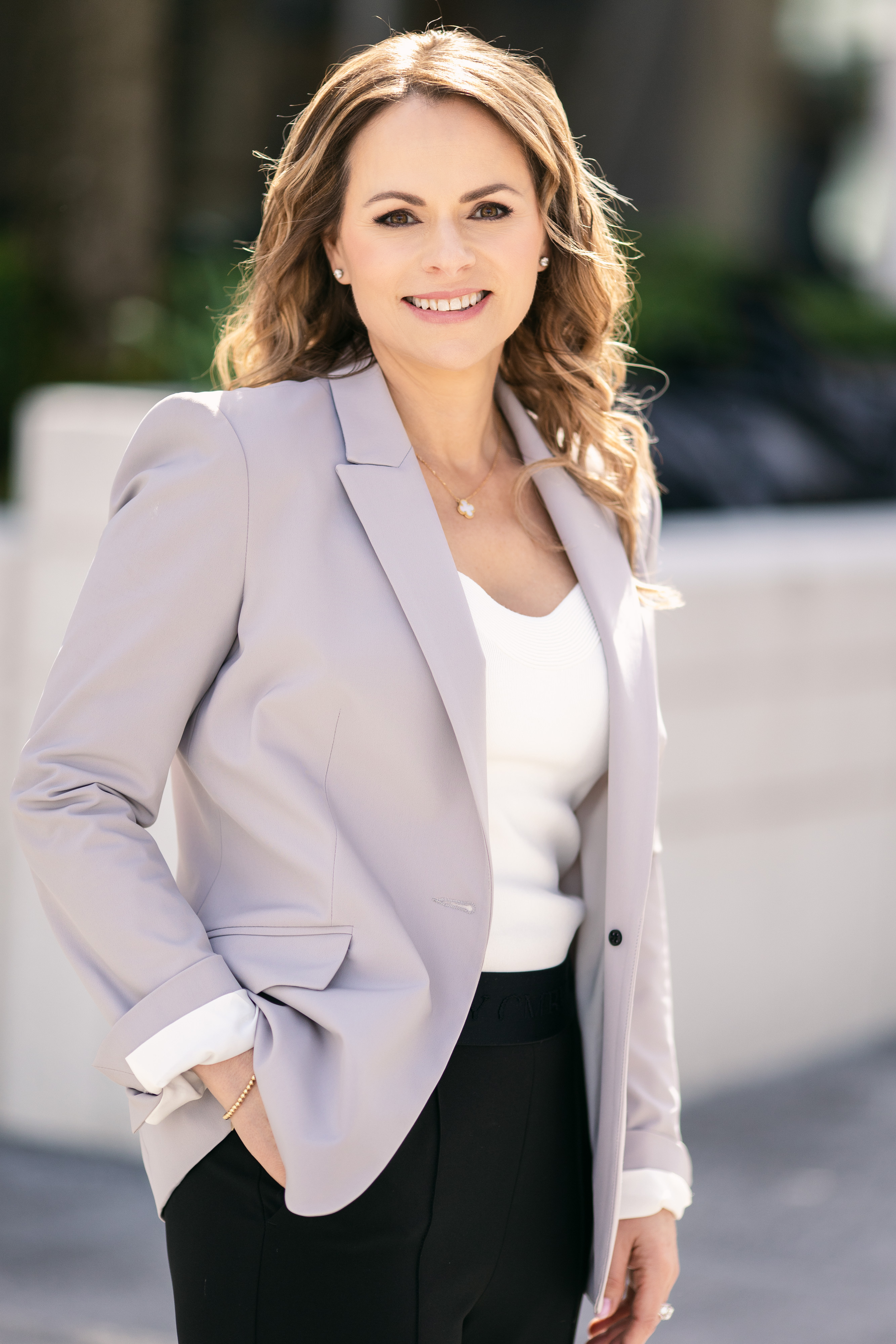 A woman with wavy brown hair smiling outdoors, wearing a light gray blazer, white top, and black pants.