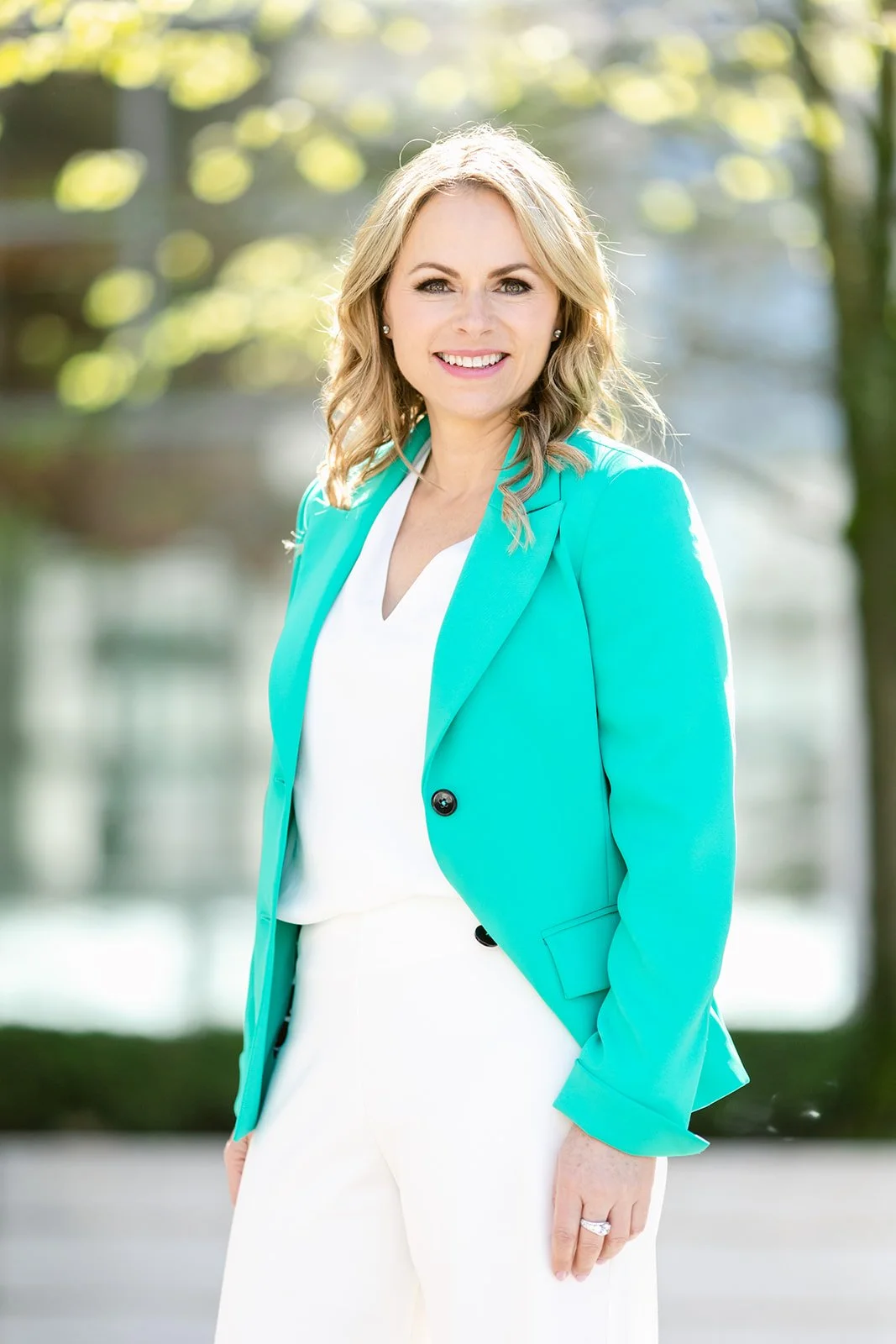 A woman with blonde hair smiling outdoors, wearing a turquoise blazer over a white blouse with white pants.