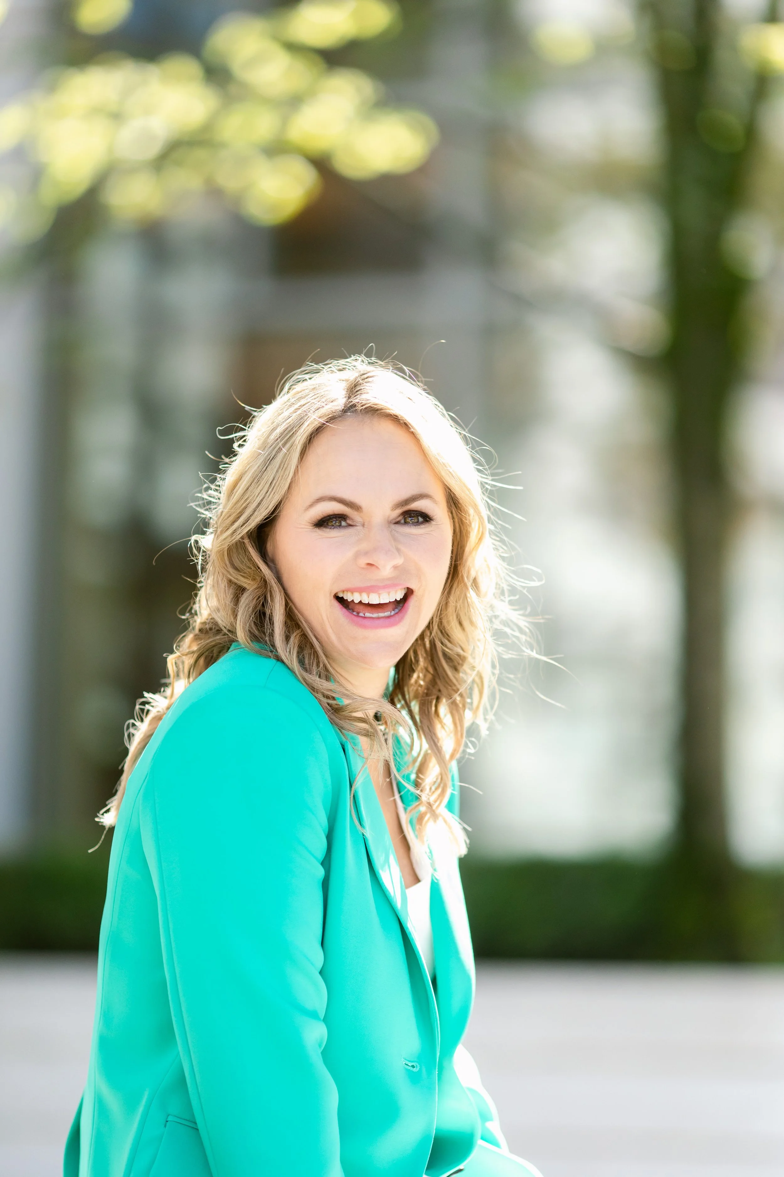 A woman with blonde hair, wearing a turquoise blazer, smiling outdoors with blurred trees and sunlight in the background.