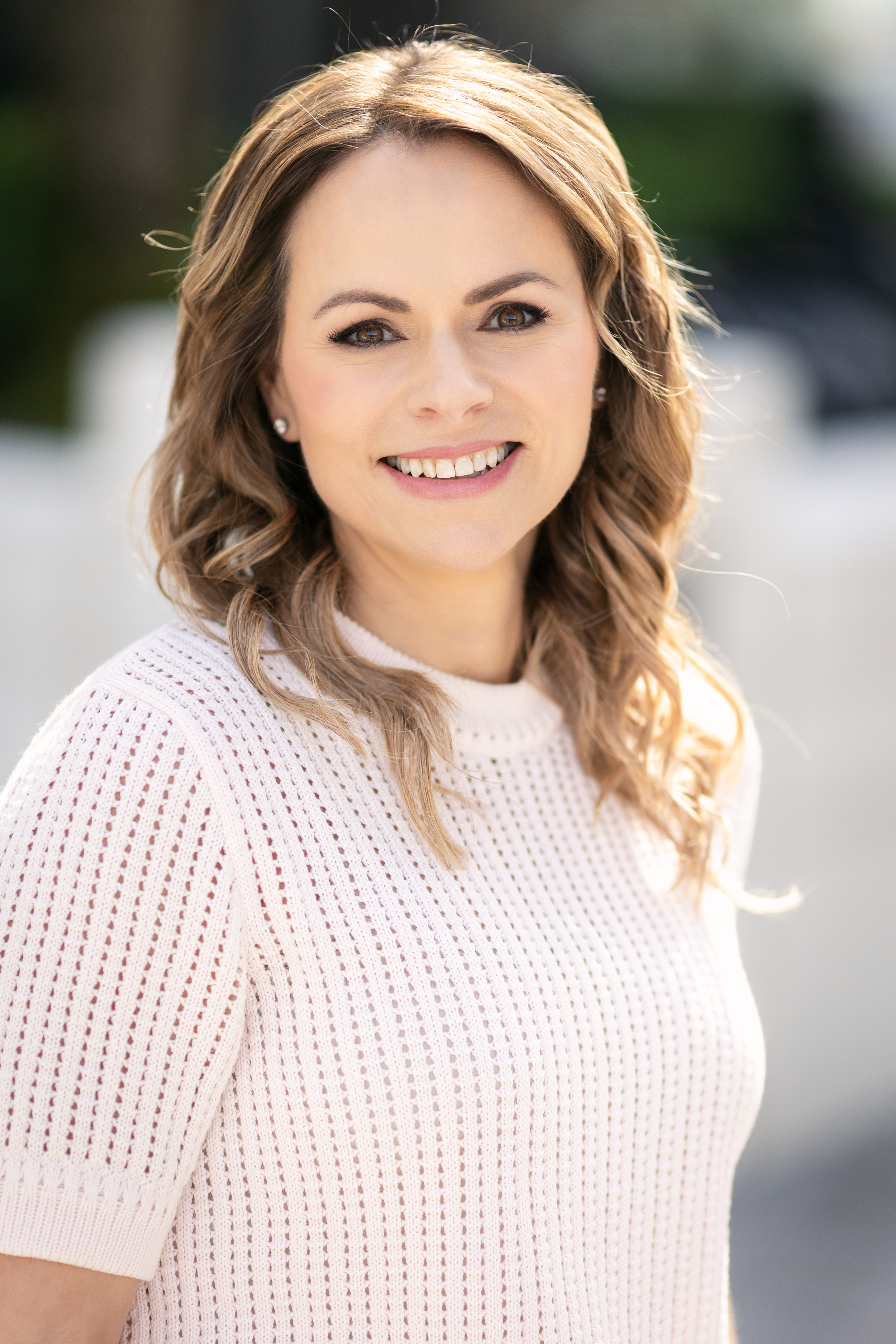 A woman with shoulder-length wavy brown hair, wearing a white knitted top, smiling outdoors in sunlight.
