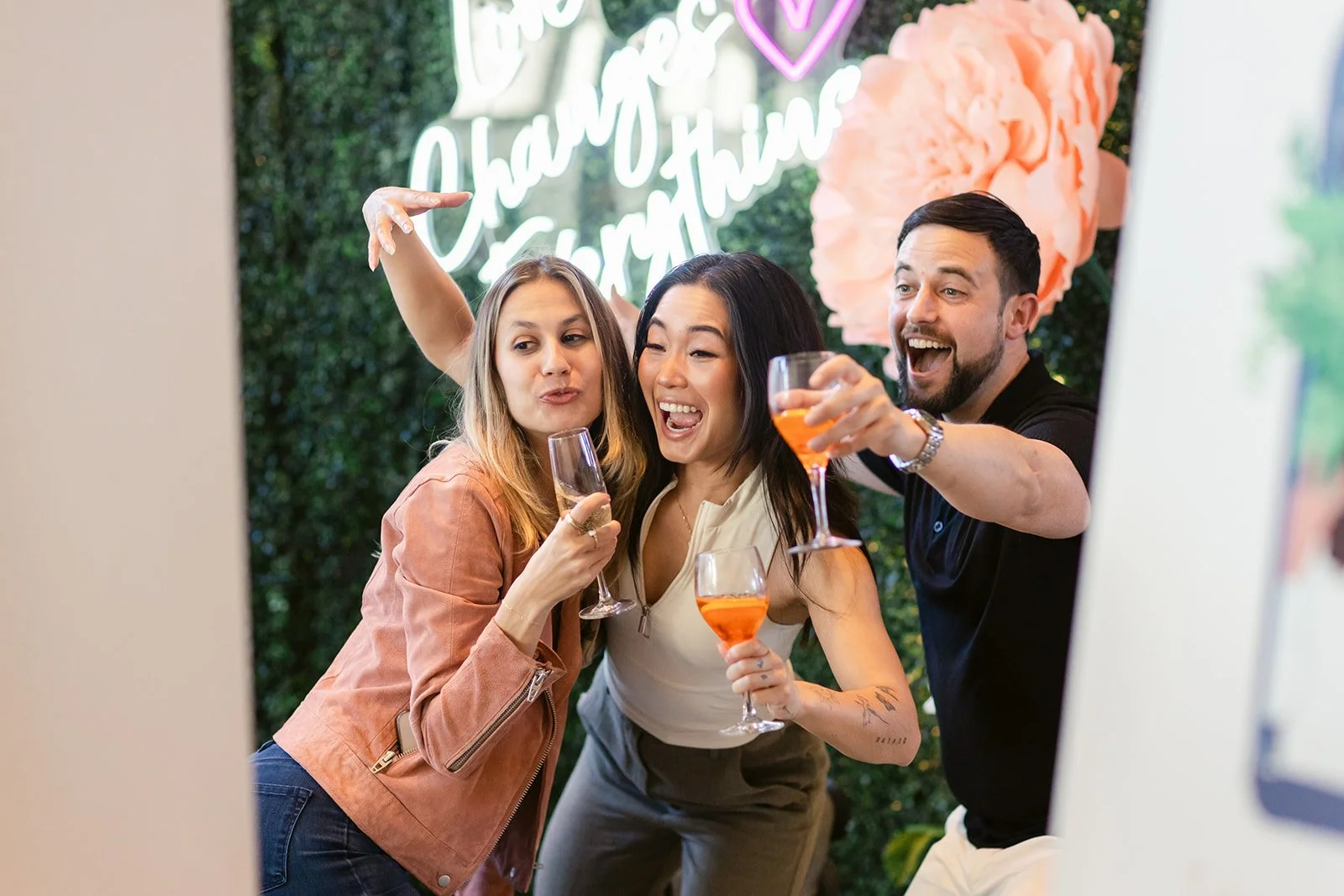 Three friends celebrating and taking a selfie at a party, holding pink-colored drinks in a decorated setting with a neon sign and pink flowers in the background.