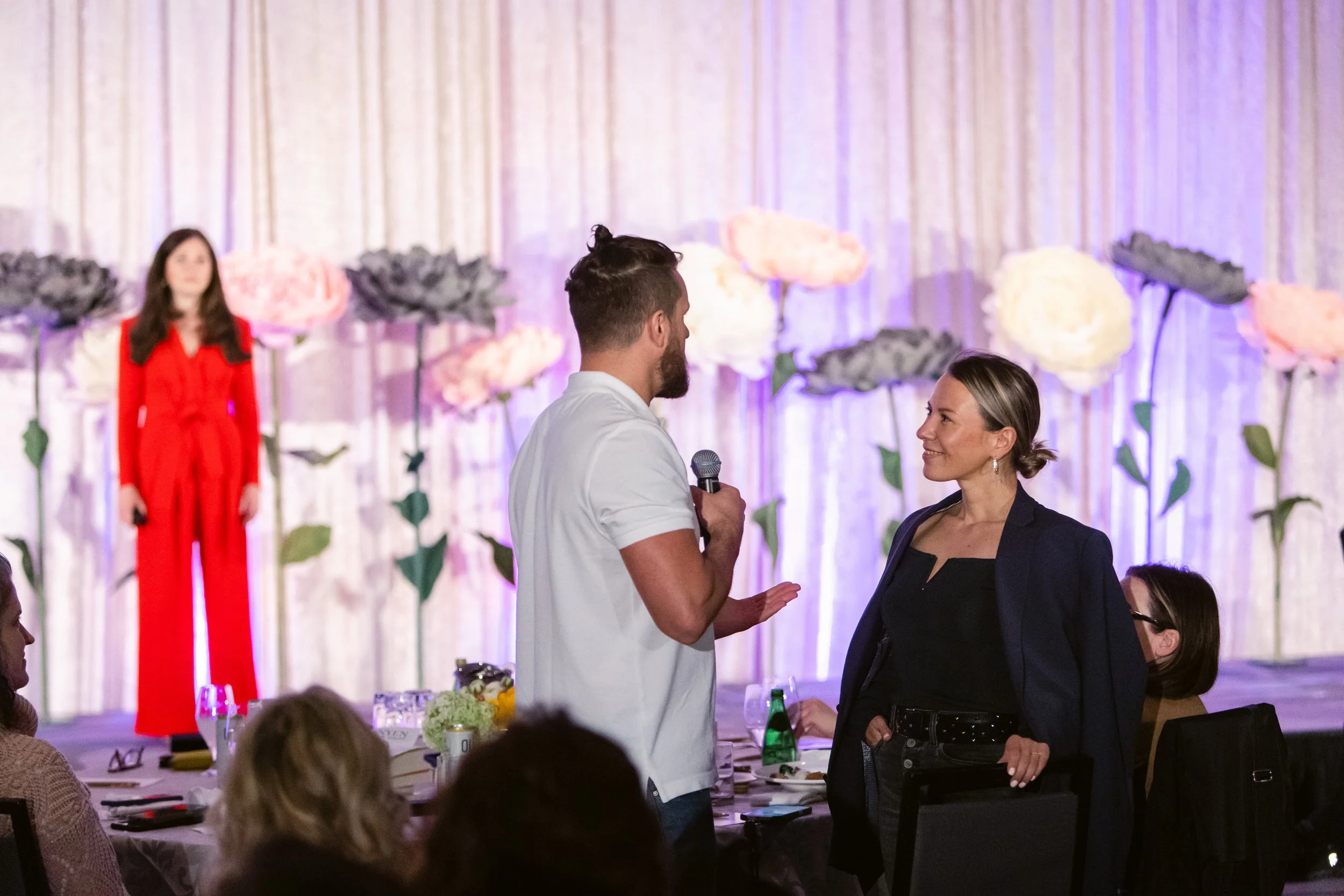 A man holding a microphone asks a woman in a black top and blazer a question or makes a comment at a formal event with floral decorations and audience members seated at tables.