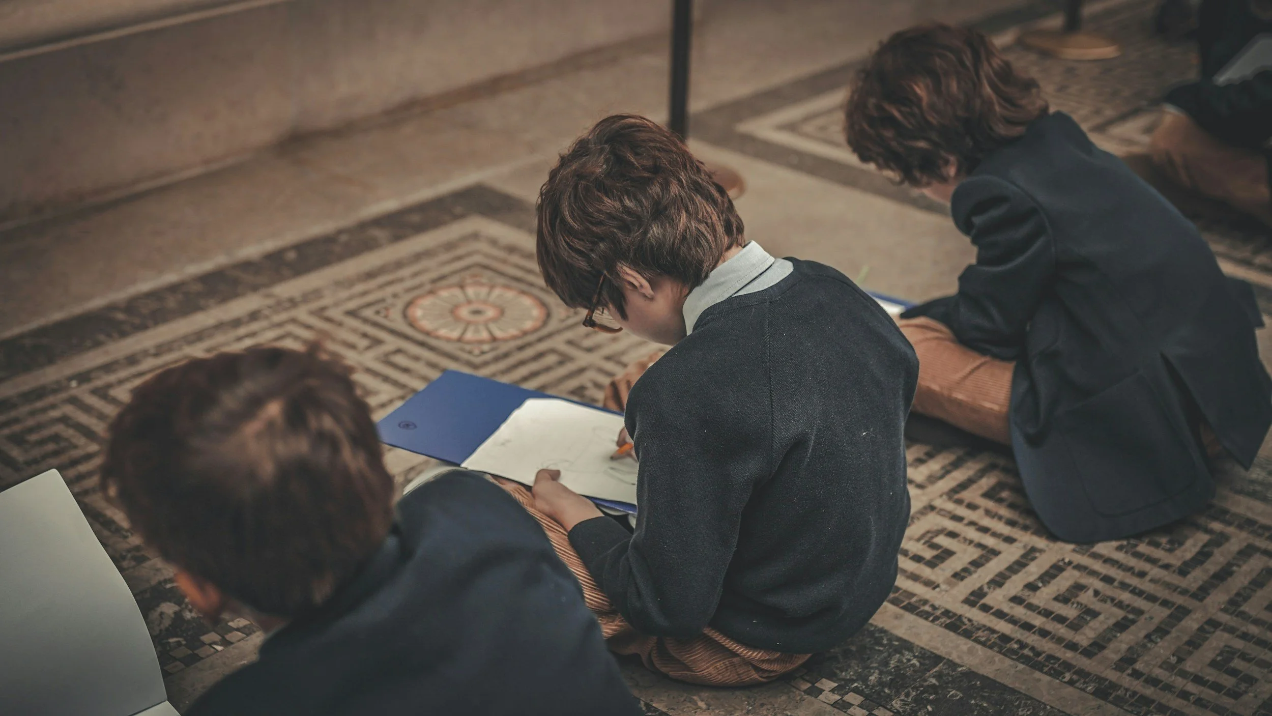 School children sitting on a patterned floor while working on notebooks.