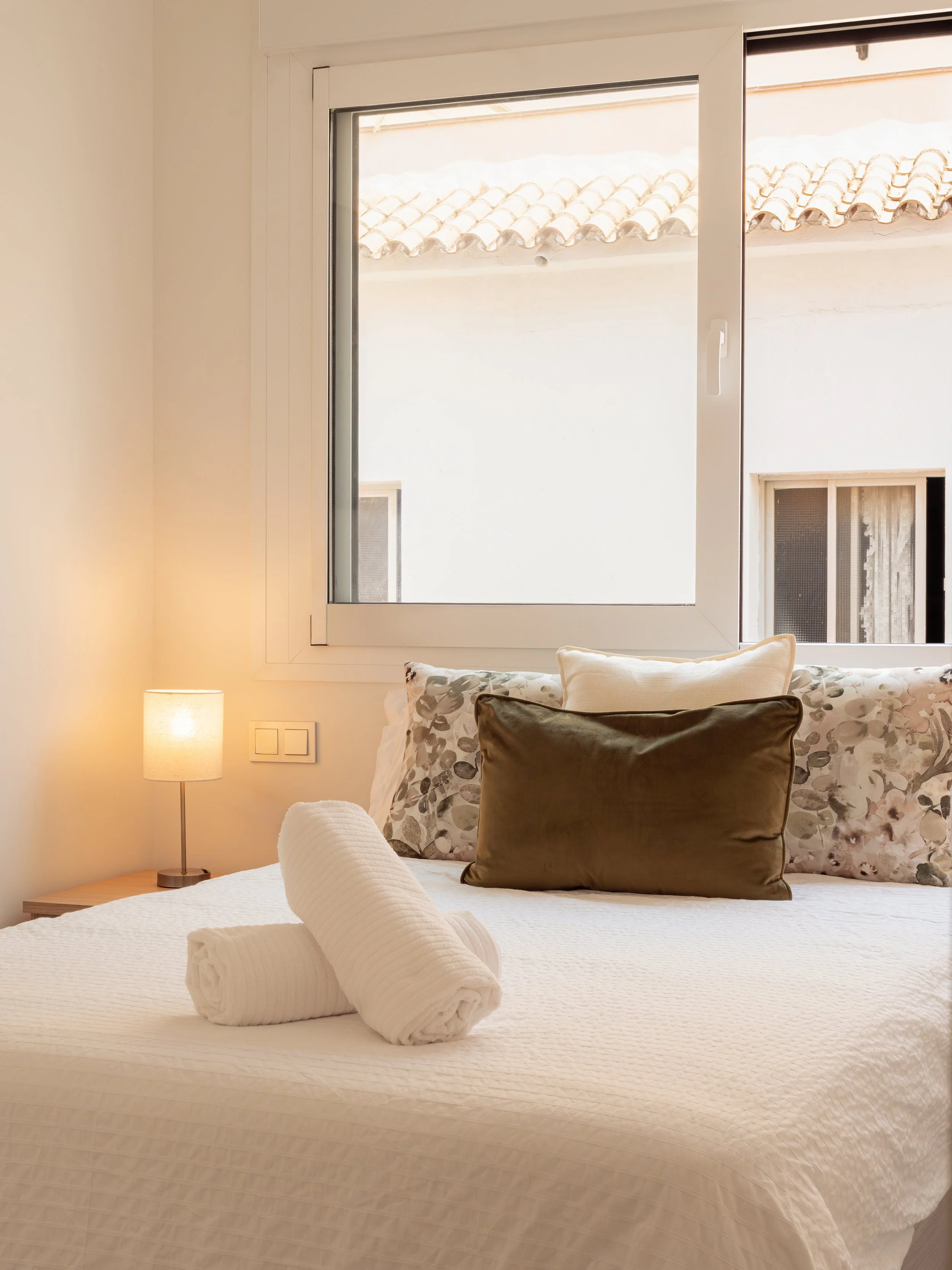 A neatly made bed with a rolled white towel at the foot, decorative pillows including a floral pattern pillow, a plain beige pillow, and a dark brown pillow. A small nightstand with a table lamp is beside the bed. There are two windows behind the bed