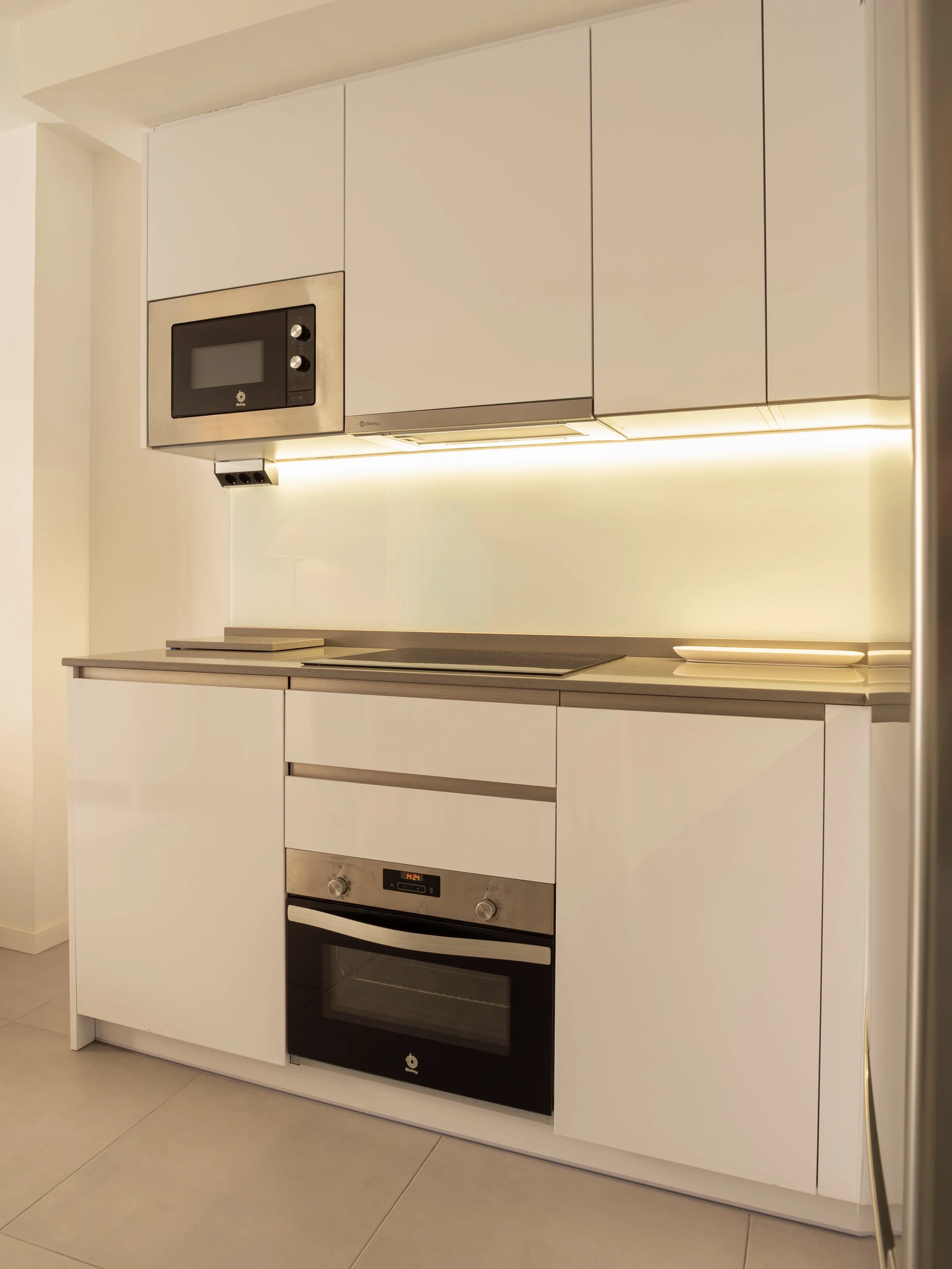 Modern kitchen with white cabinets and built-in appliances, including a microwave, oven, and stovetop, with under-cabinet lighting.