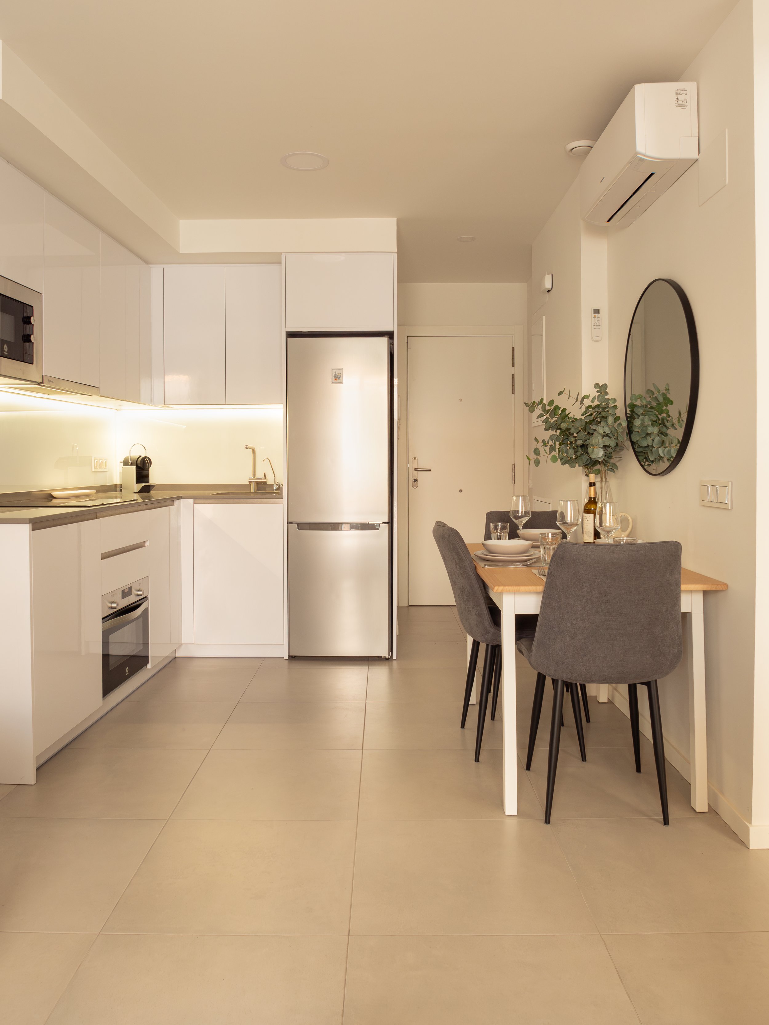 Modern open-plan kitchen and dining area with white cabinetry, stainless steel refrigerator, and a small dining table with four gray chairs. Decor includes a large round mirror, green plant, and tableware. The room has beige tiled flooring and neutra