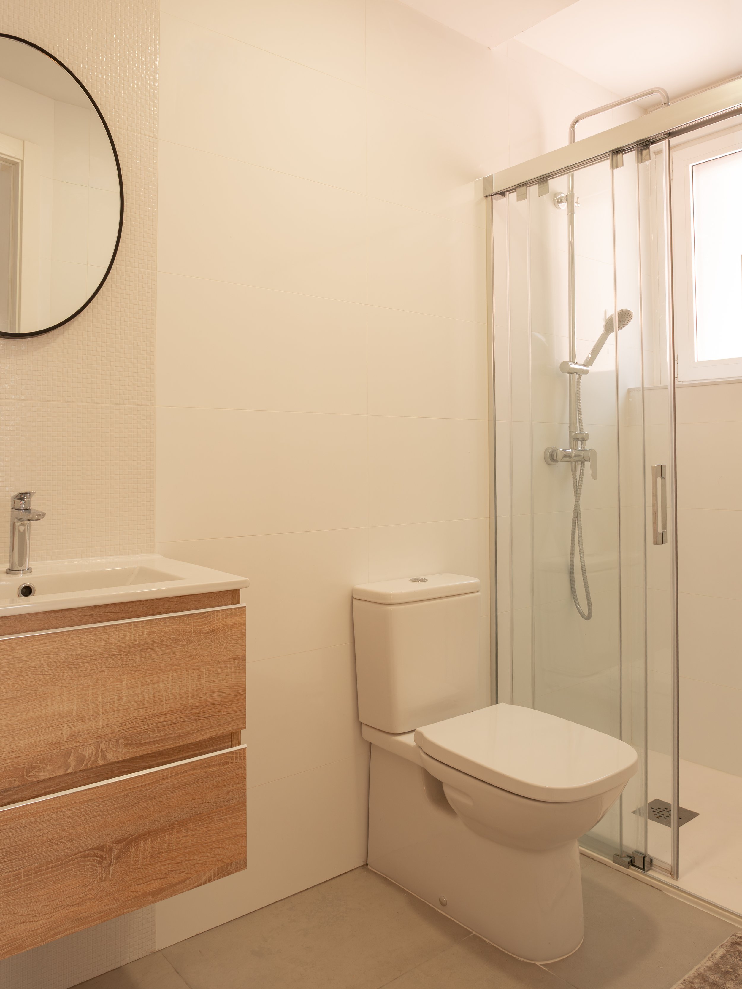 Modern bathroom with a white toilet, wooden vanity with a white sink, a round mirror, and a glass shower enclosure with a rainfall showerhead.