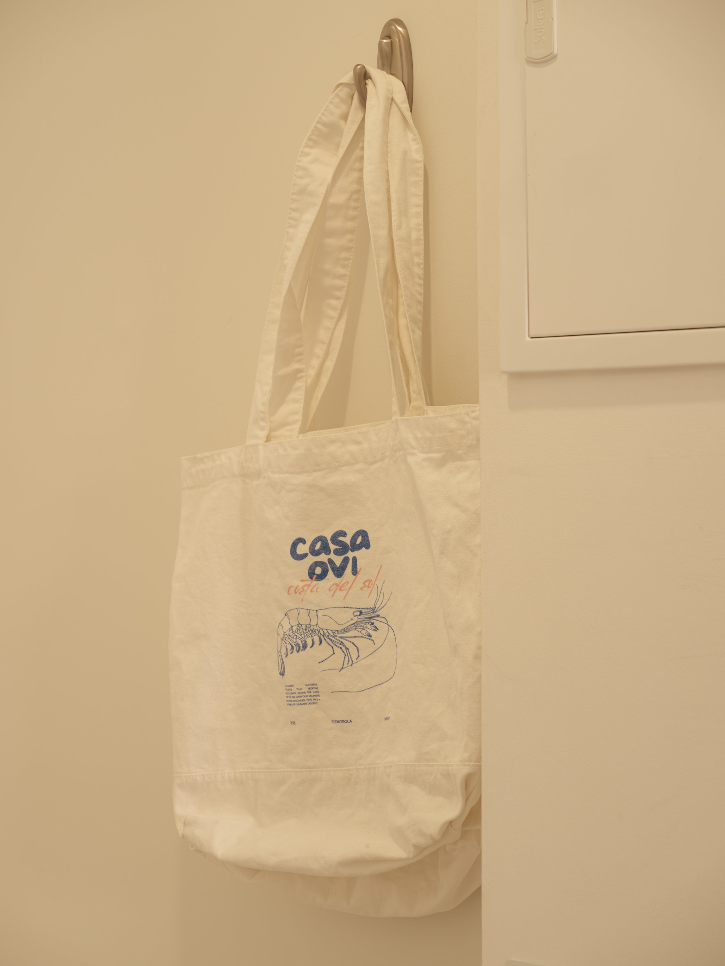 A white tote bag hanging on a door handle with the logo 'casa ovi' and a drawing of a lobster.