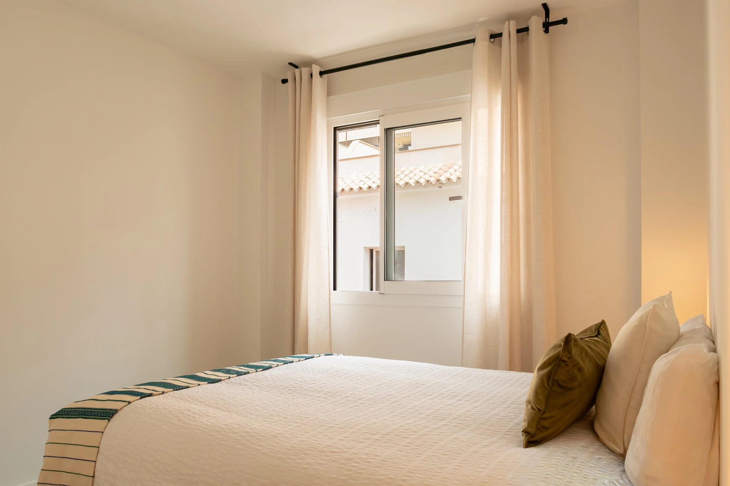 View of a bedroom with a white bedspread, beige pillows, and a window with light curtains, outside rooftops are visible.