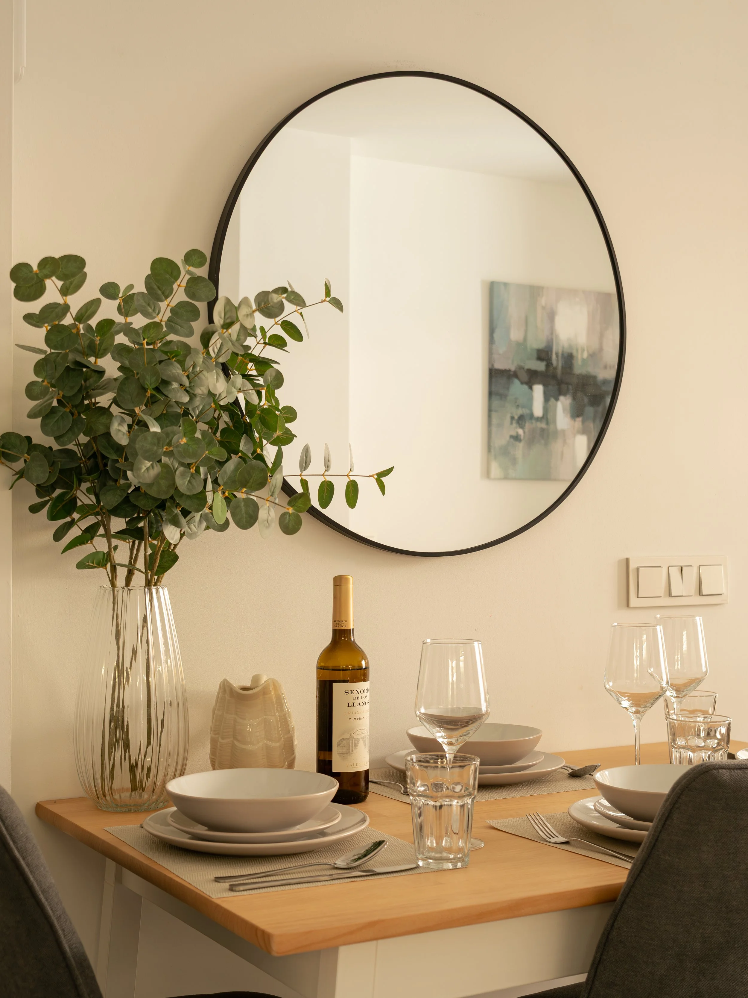 A dining table set with white plates, glasses, and cutlery, with a large vase of green leafy branches and a bottle of white wine against a wall with a round mirror and a painting.