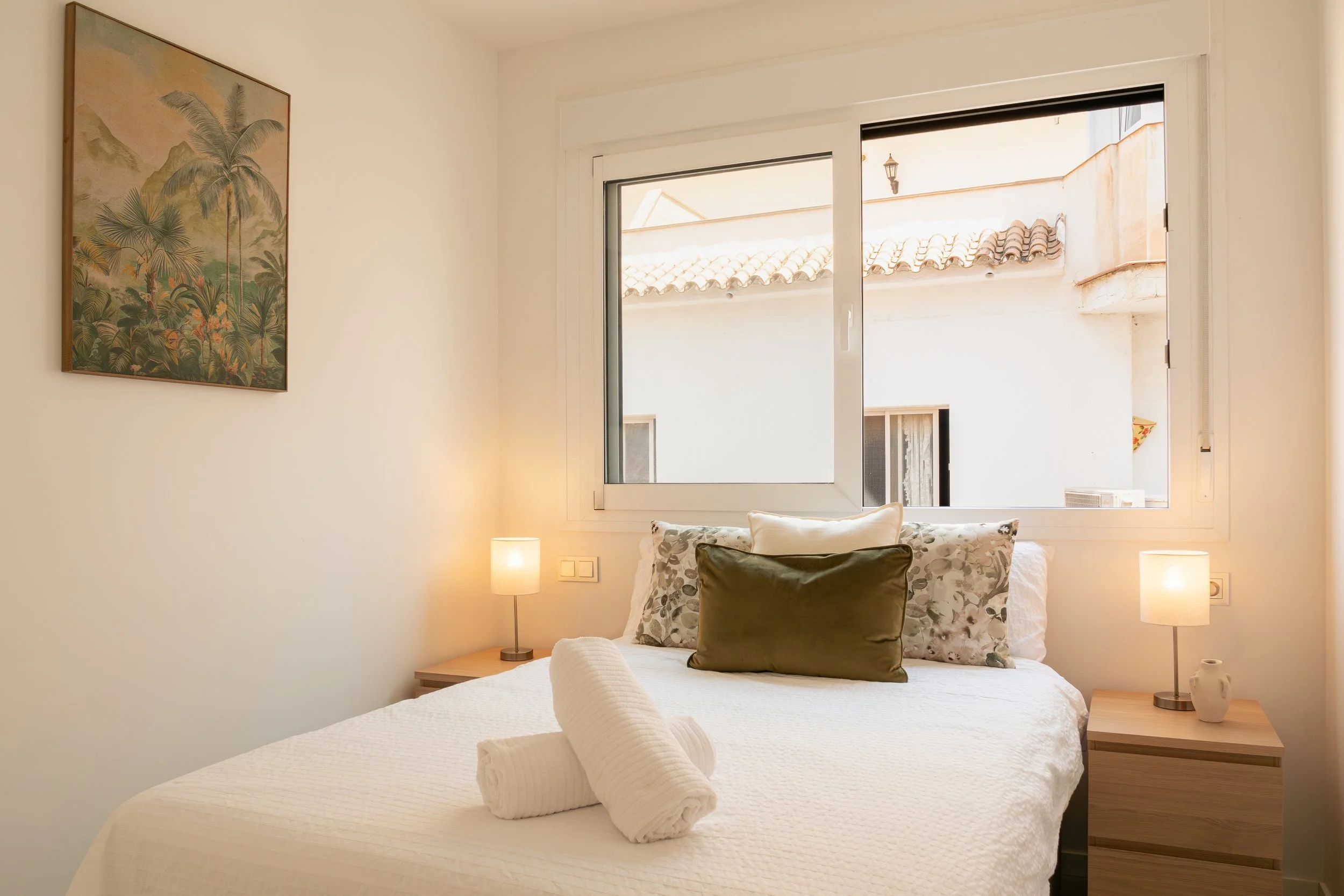 A bedroom with a large bed covered in white bedding, decorated with rolled towels and throw pillows. There are two bedside tables with lamps, and a window behind the bed shows an outdoor view of tiled rooftops and a wall with a lantern. A framed trop