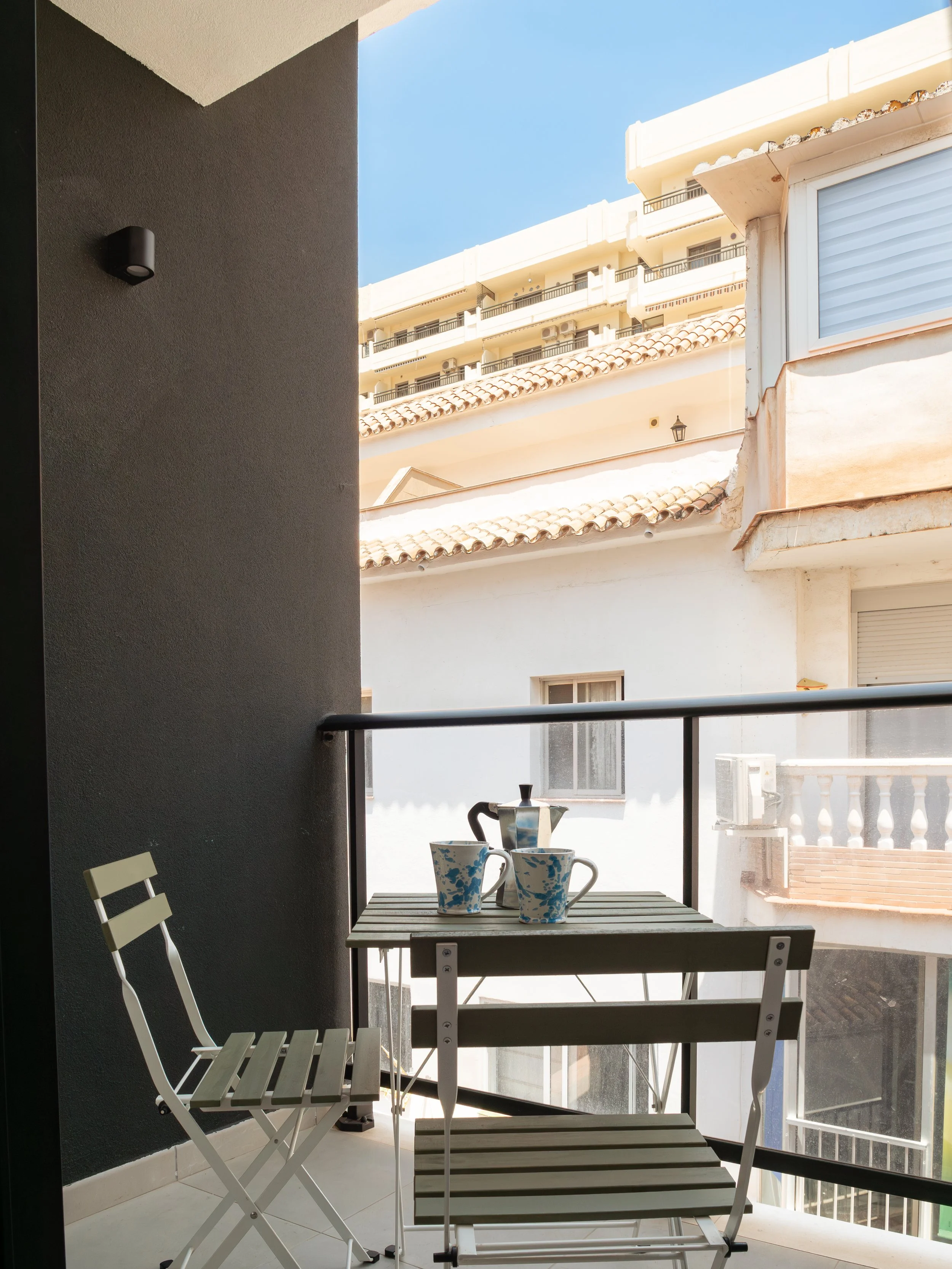 Balcony view with a small table holding four mugs, two matching white chairs, overlooking neighboring buildings with balconies and a blue sky.