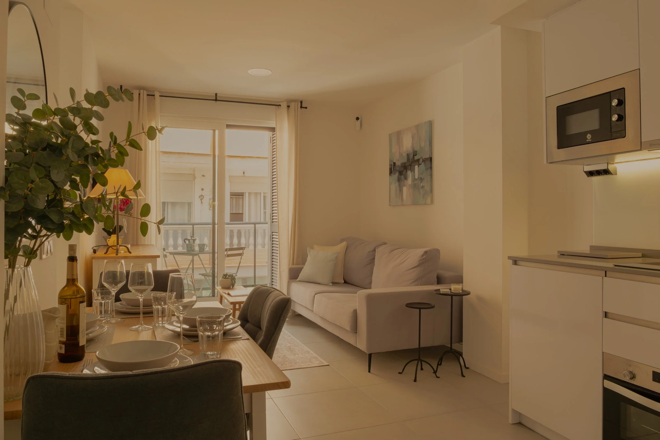 A living room with a beige sofa, abstract wall art, a dining table set with wine glasses and dishes, and a balcony with chairs outside. The room has light-colored walls and curtains, and a kitchen on the right side.