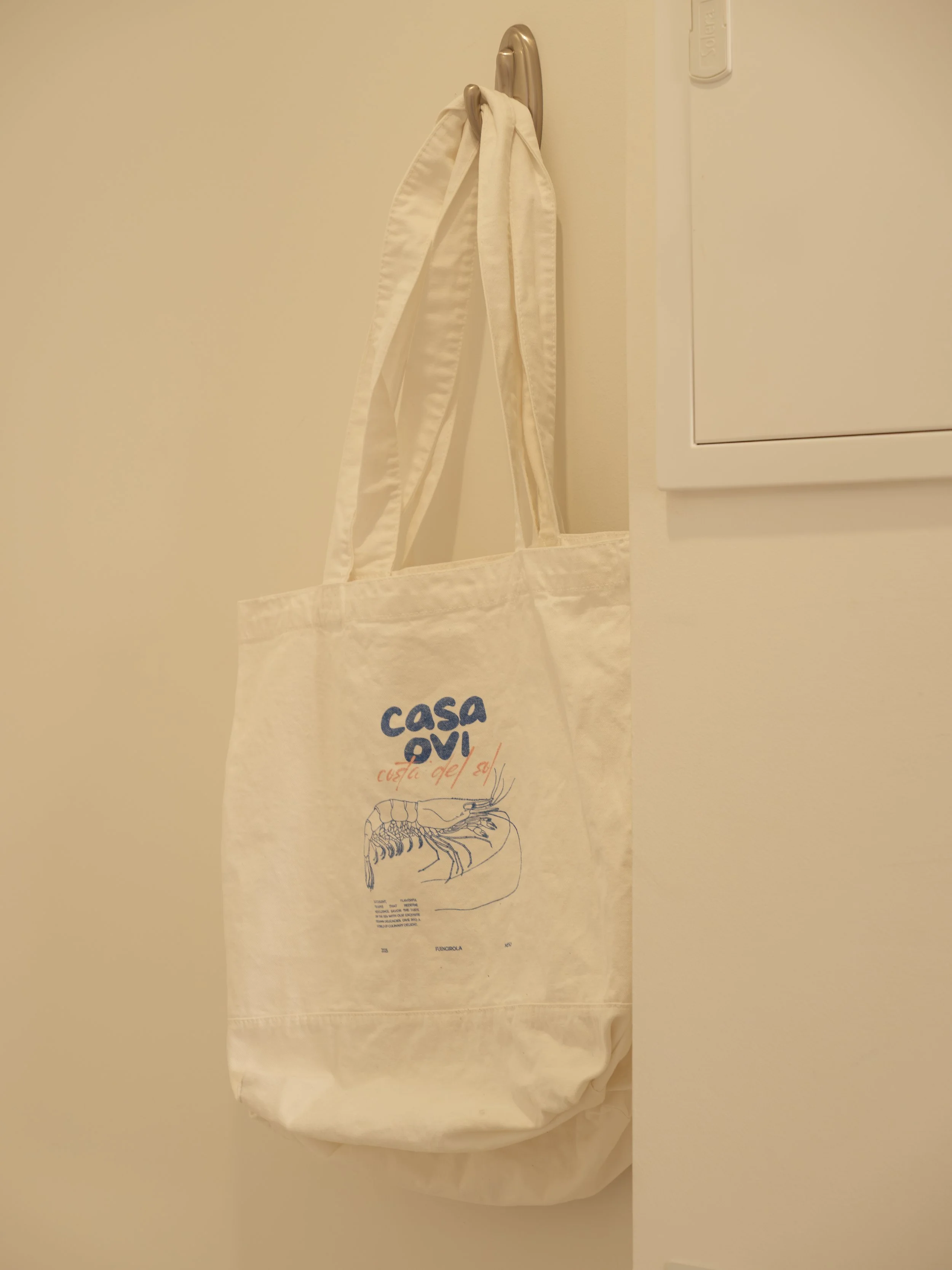 A white reusable shopping bag hanging on a beige door hook. The bag has a blue logo and a lobster illustration on it, with text that says "Casa Ovi" and some other smaller text.
